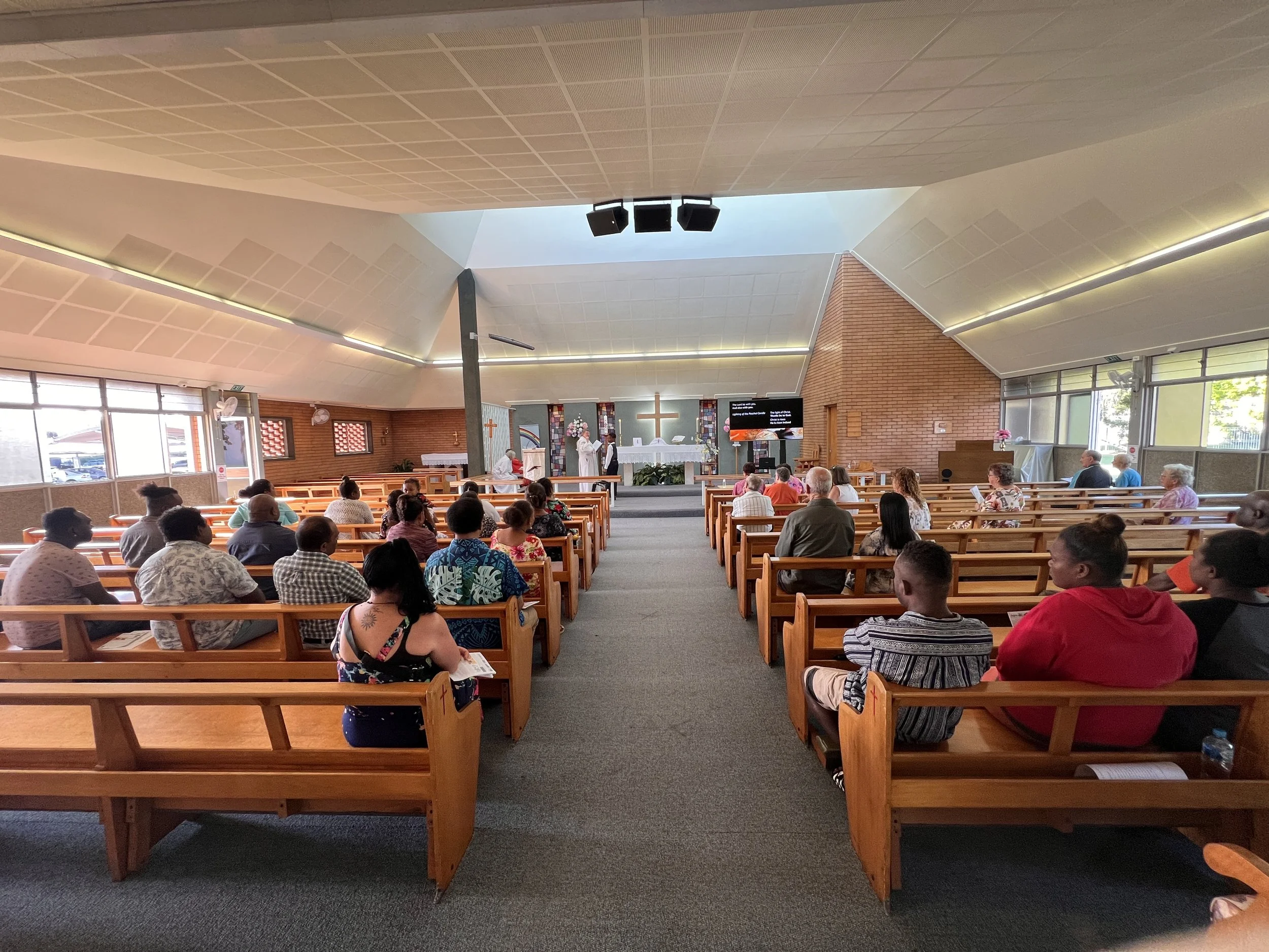 Interior of a church with wooden pews filled with people attending a service. The altar at the front has a cross, flowers, and a priest dressed in white robes. Large windows and modern ceiling design are visible.