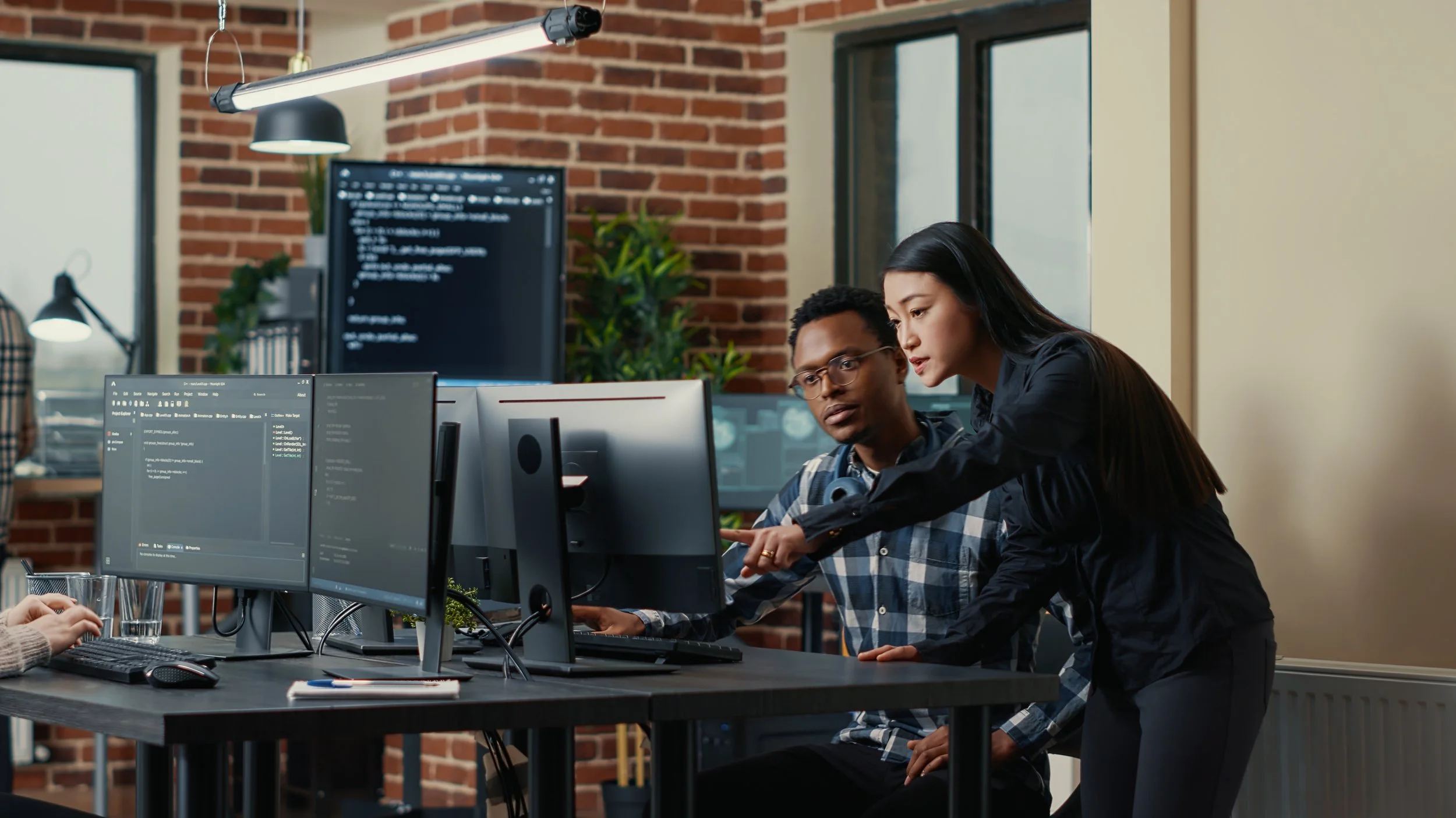 Two people working on computer screens in an office; one person is seated and the other is standing, pointing at the screen.