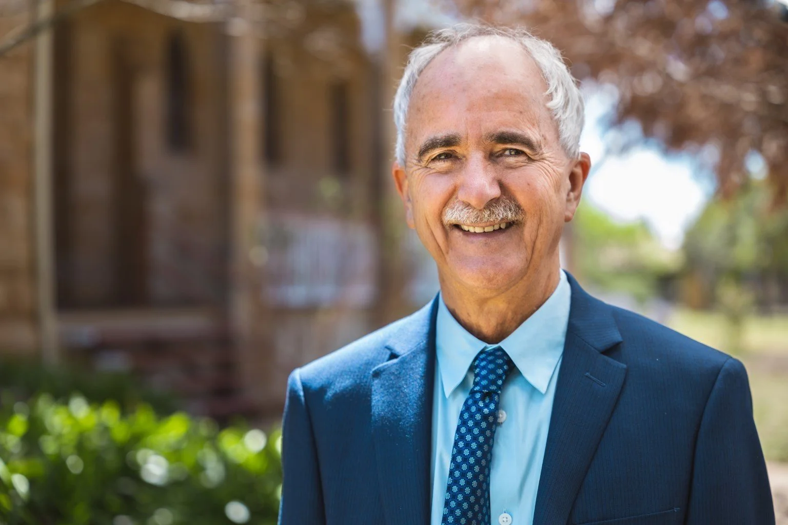 Smiling elderly man in a blue suit and patterned tie outside with trees in the background.