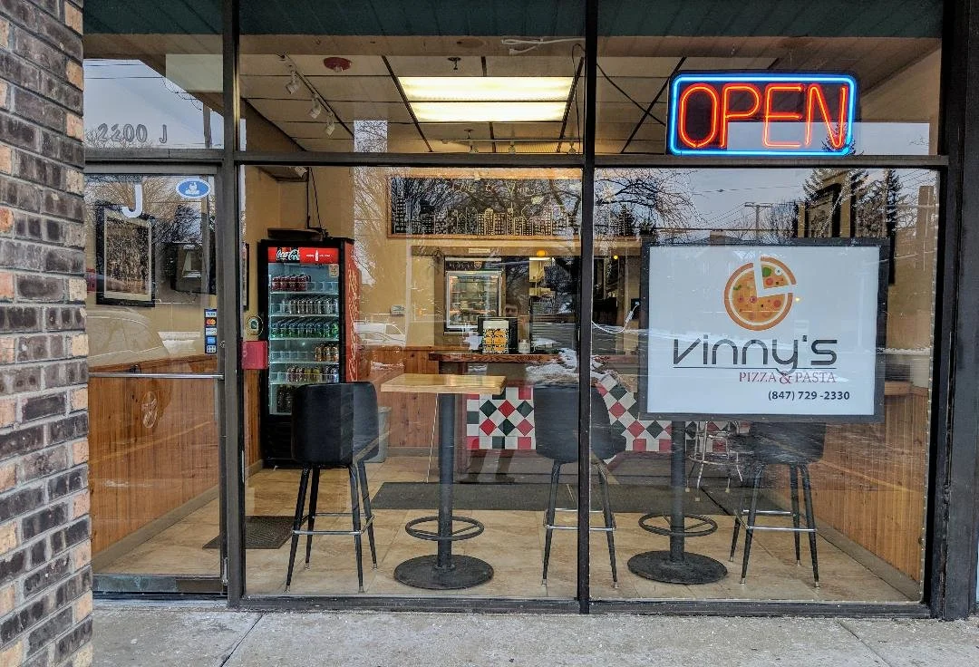 View of the interior of Vinny's Pizza & Pasta restaurant through a glass storefront door and windows, featuring a neon 'Open' sign, a Coca-Cola vending machine, three black bar chairs around a high table, and a large sign displaying the restaurant's name and contact information.