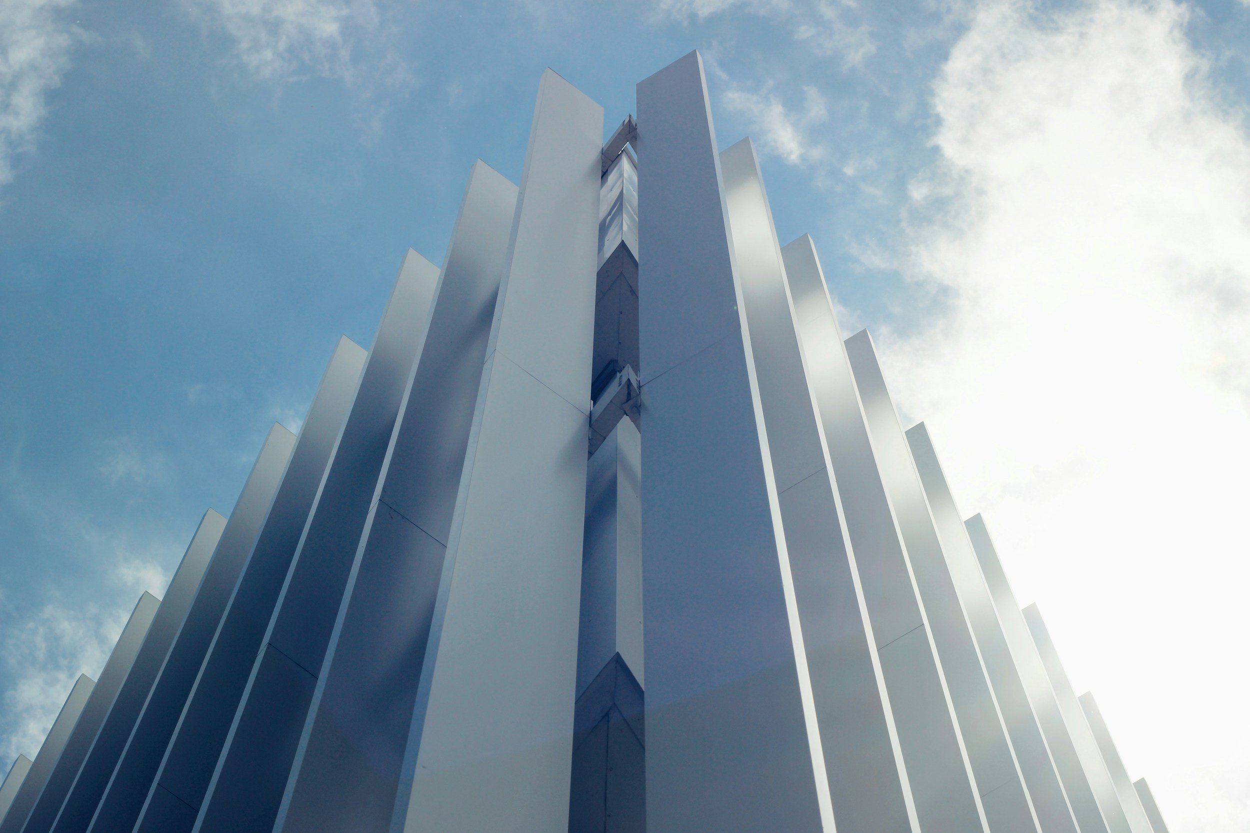 A modern high-rise building with a sleek, white facade and vertical panels, seen from below against a partly cloudy sky with sunlight.