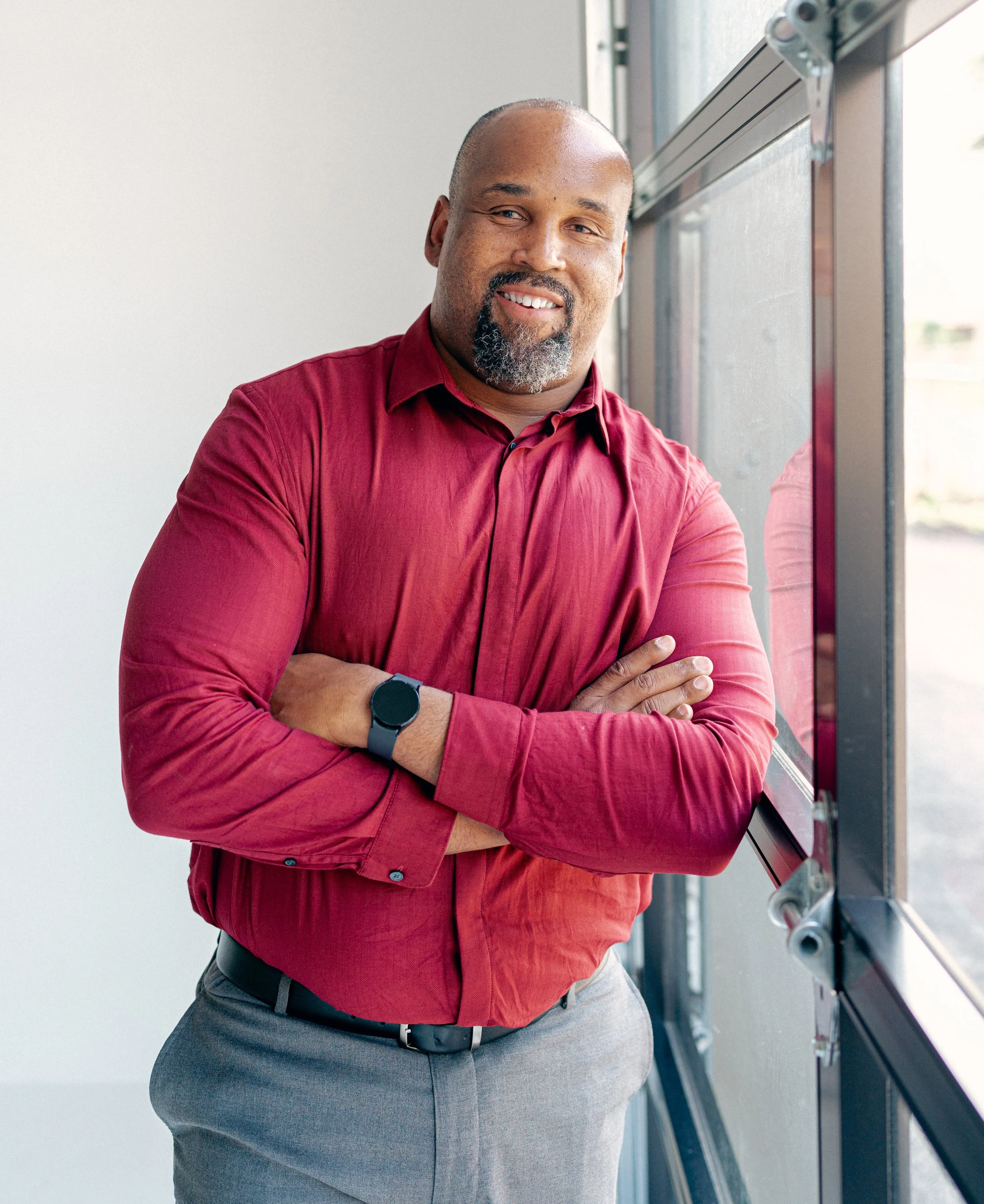 A confident man standing next to a window with arms crossed, dressed in a red shirt and gray pants, smiling.