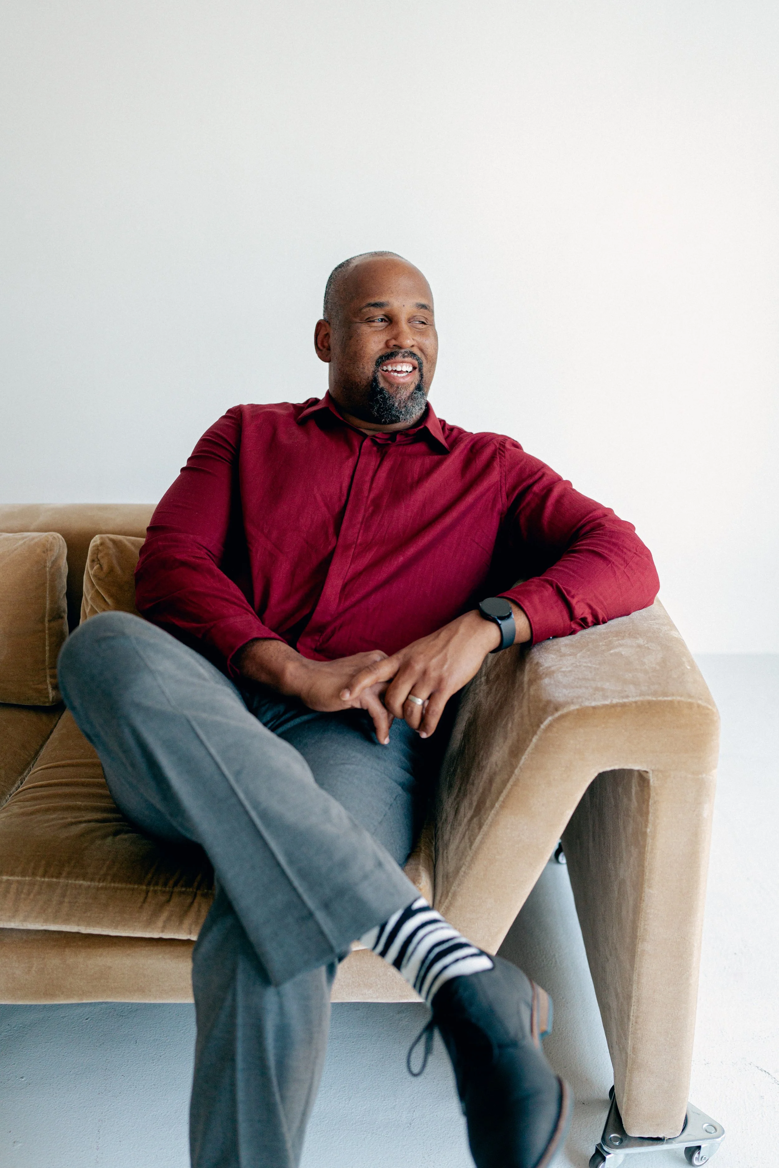 A man sitting on a beige sofa with a relaxed posture, smiling, wearing a red shirt, gray pants, striped socks, and black shoes, with a watch on his wrist, in a minimalistic room with white walls.