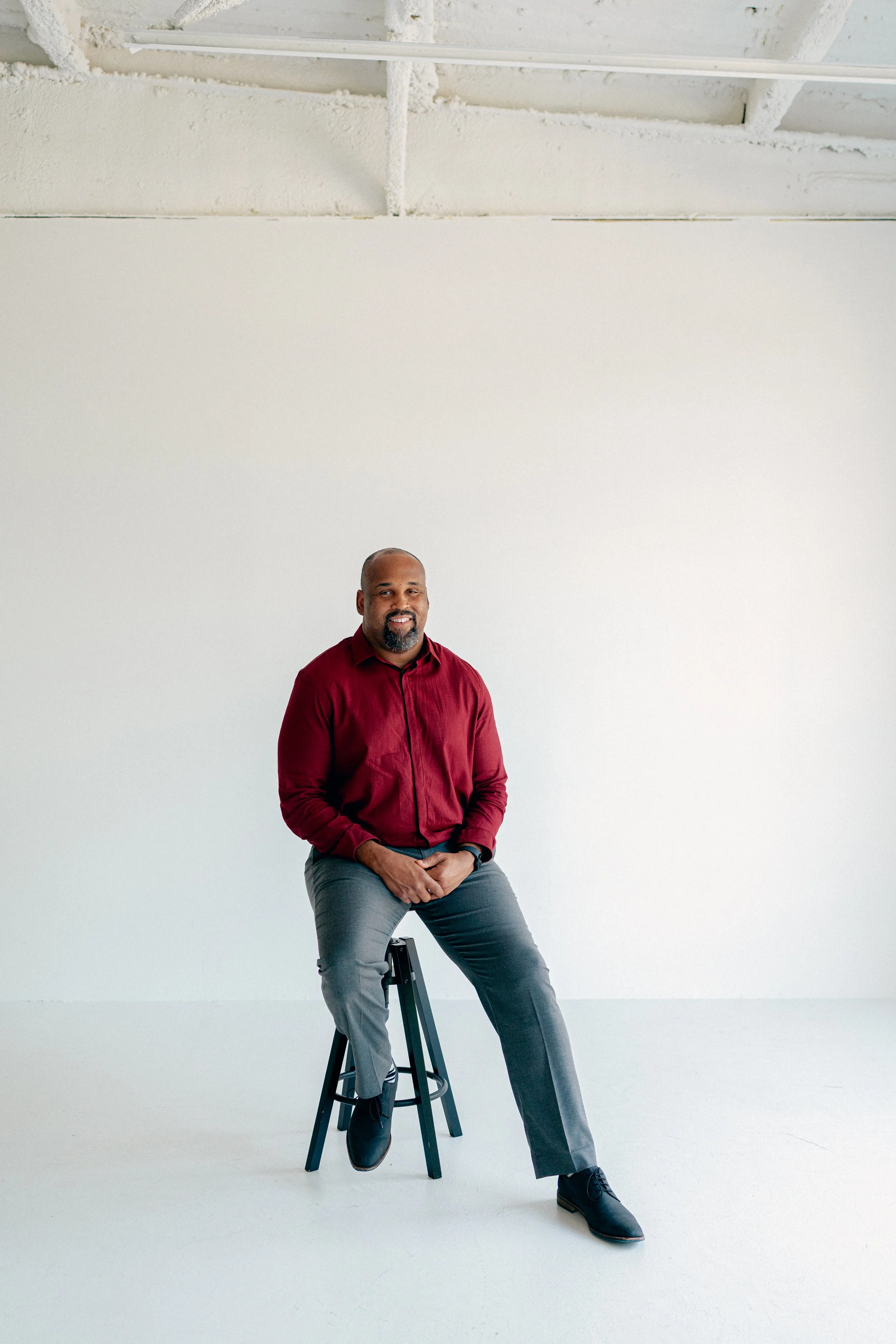 A man with a beard wearing a red shirt and gray pants sitting on a black high stool against a white background.