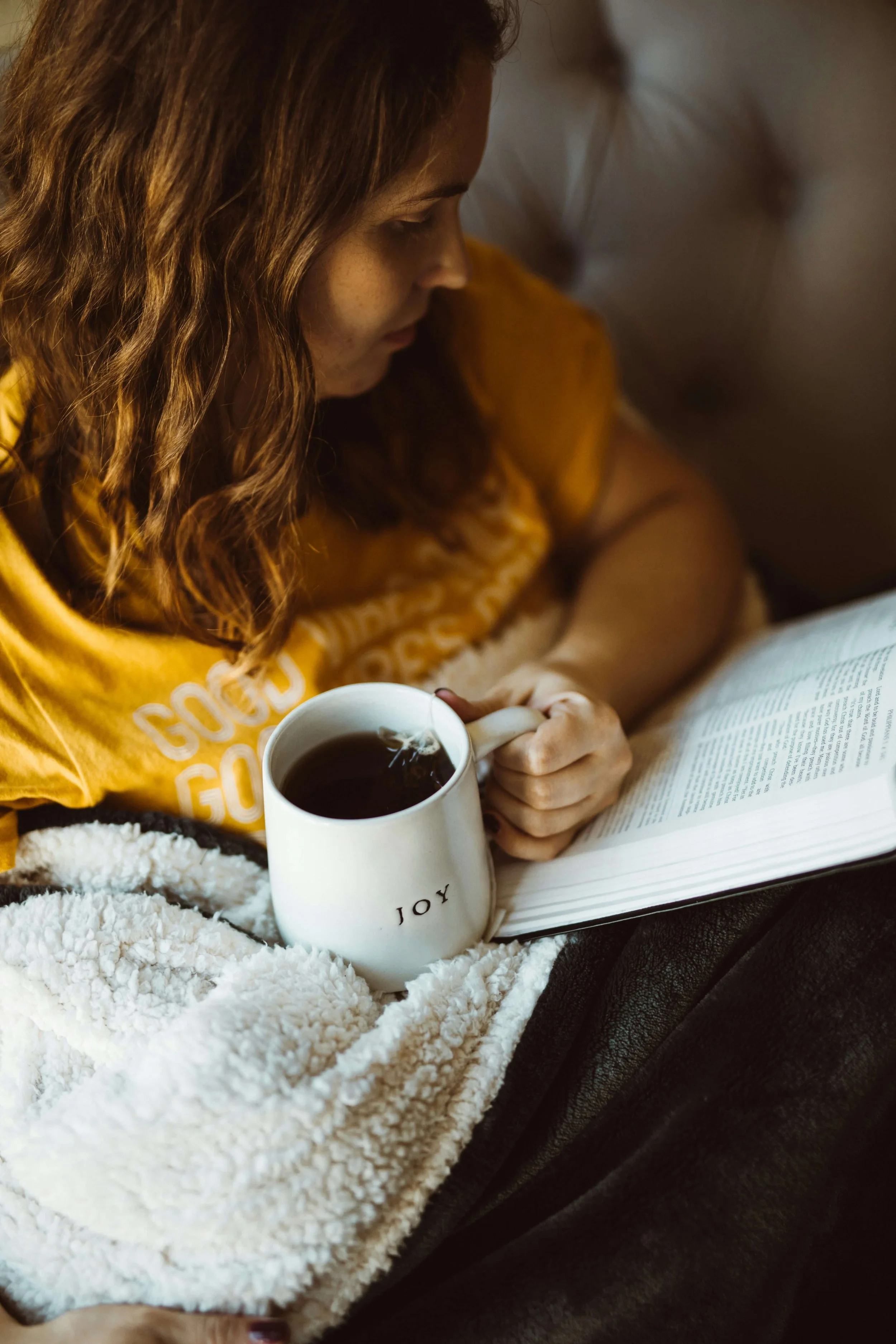 Woman sitting under a blanket reading a book and holding a mug of tea in a cozy bedroom.