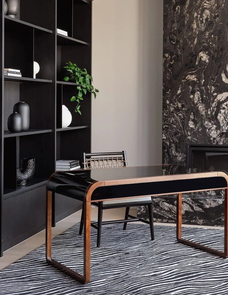 Modern office interior with a black desk featuring wooden edges, a black chair with a woven backrest, black and white striped rug, and a black wall shelf with decorative items and a plant. Black marble wall on the right side.