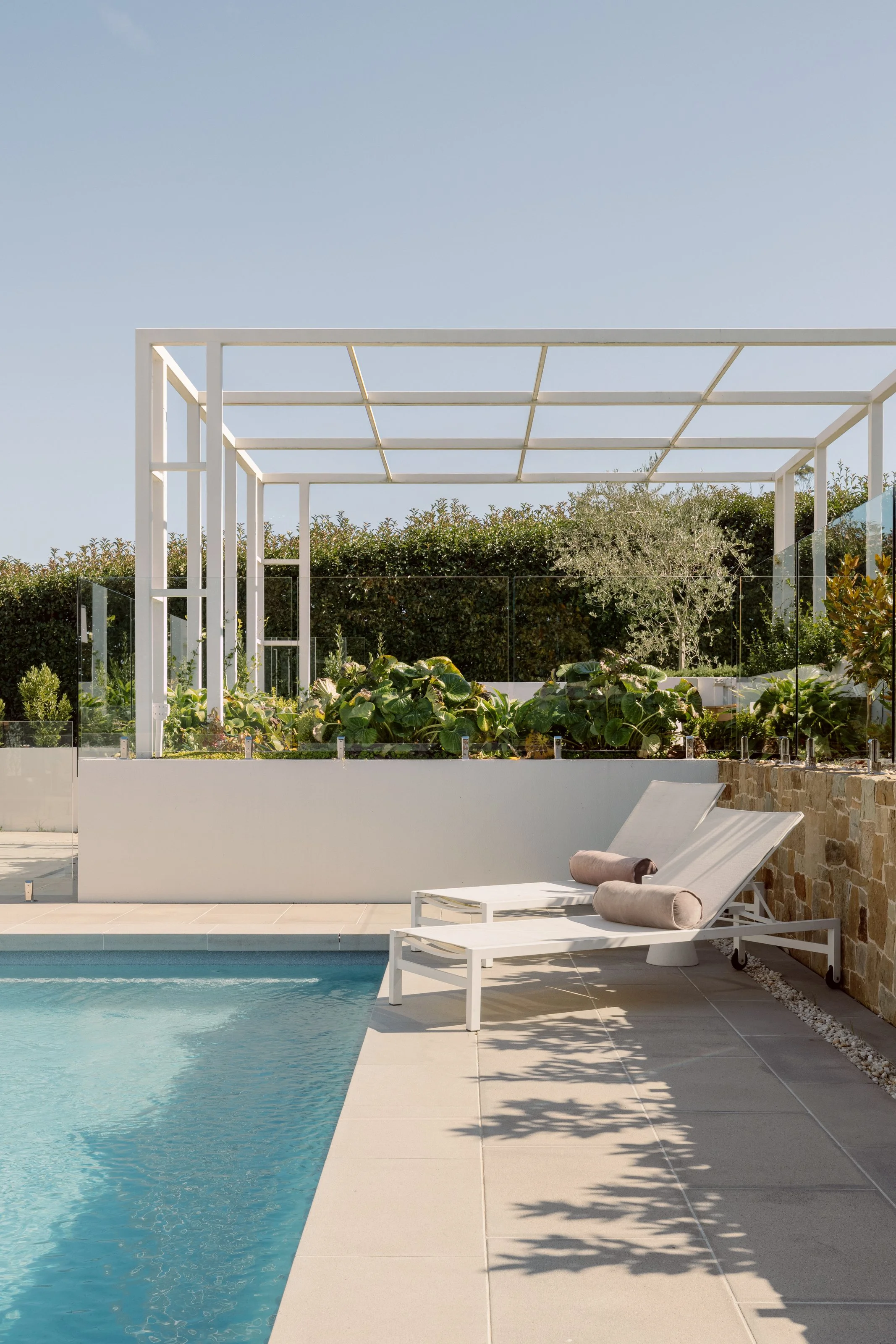 Modern backyard patio with a swimming pool, two white lounge chairs with beige cushions, and a garden with lush green plants and trees in the background.