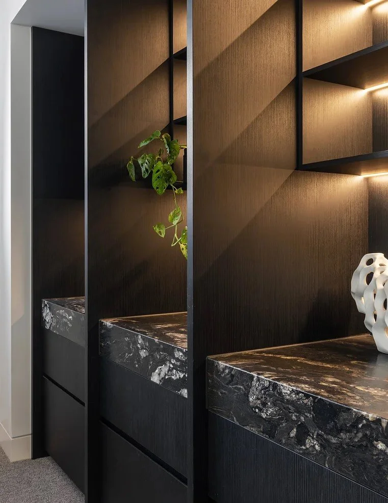 Modern interior wall with black wood shelving units, illuminated by built-in lights, featuring a marble countertop, a small green plant, and a decorative white sculpture.