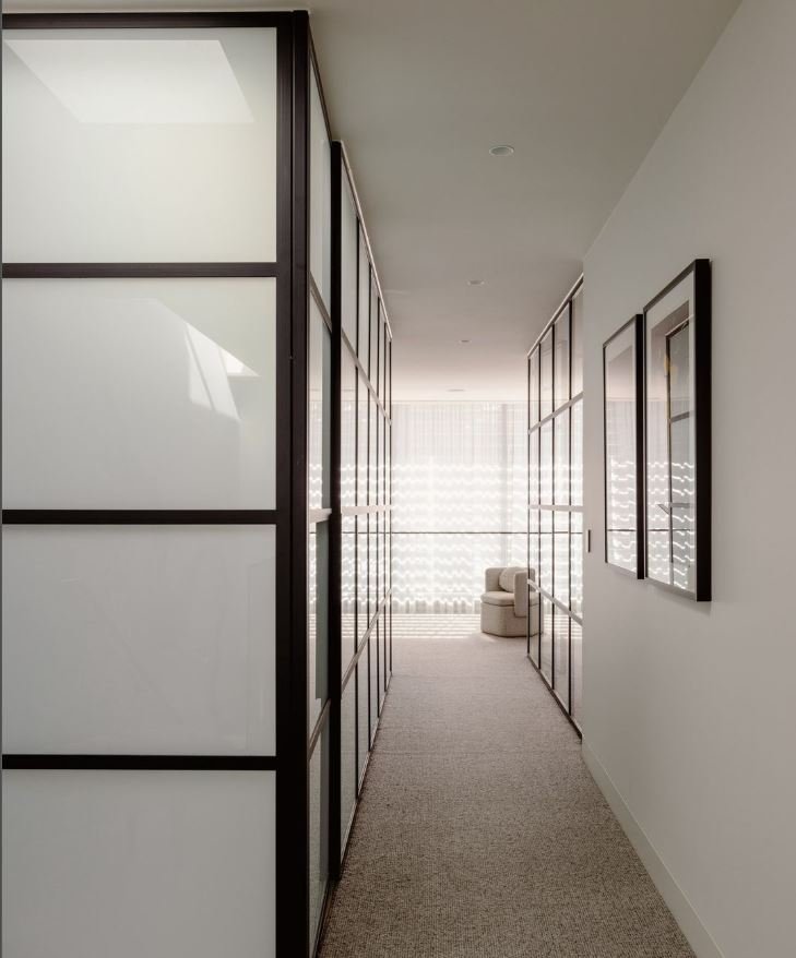 A modern, minimalist hallway with frosted glass doors on both sides and a small corner seating area at the end with sheer curtains allowing natural light to fill the space.