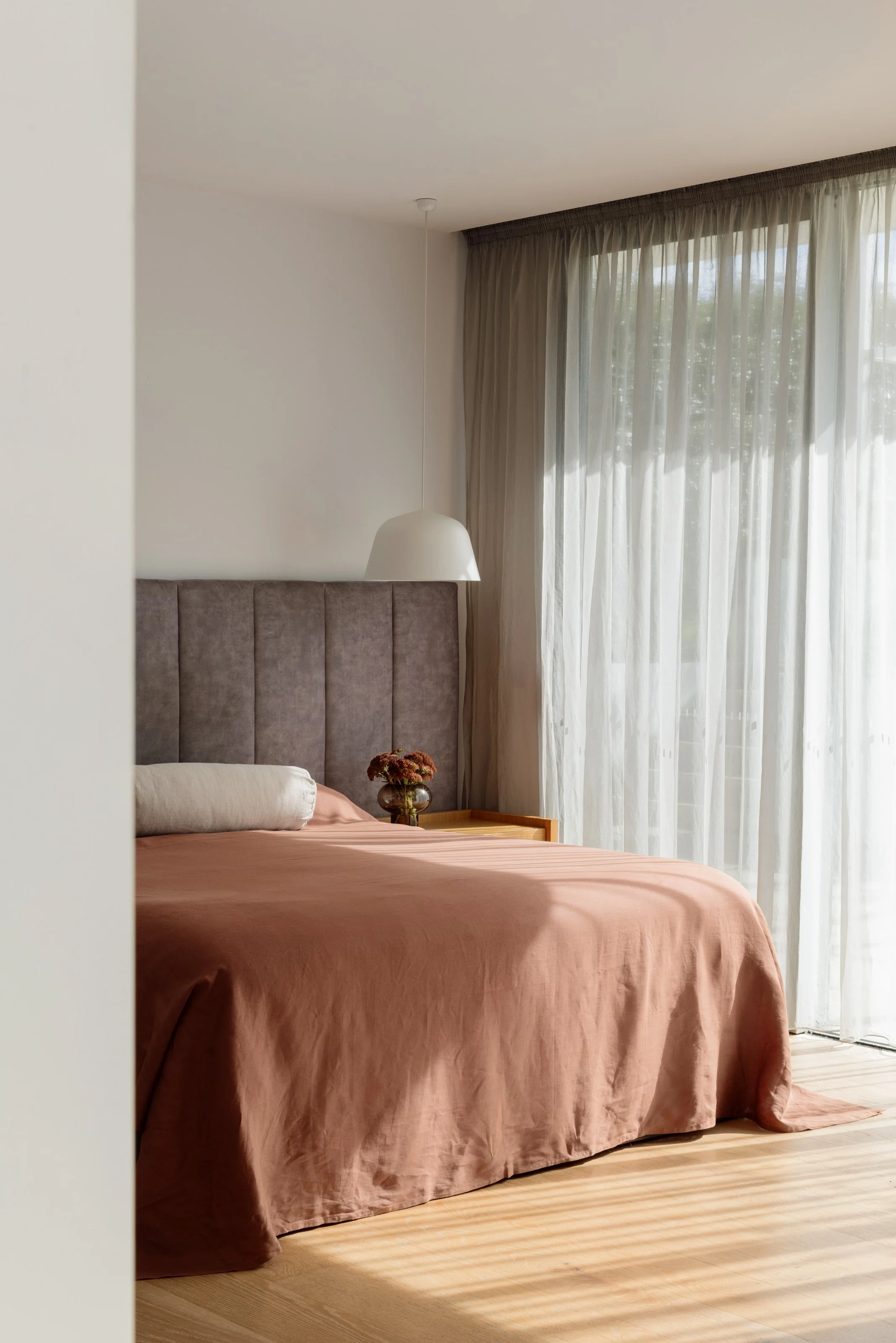 A neatly made bed with a dusty pink bedspread, a long white pillow, and a brown velvet headboard in a bright bedroom with sheer curtains and sunlight streaming in.