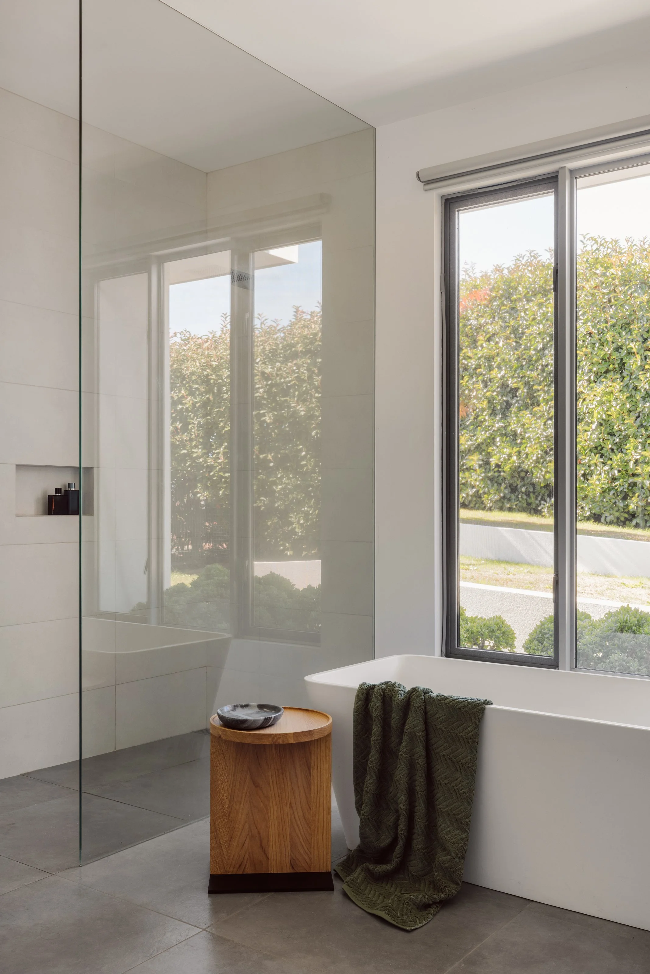 Modern bathroom with bathtub, wooden side table, dark green towel, large window with outside greenery, and minimalist design.