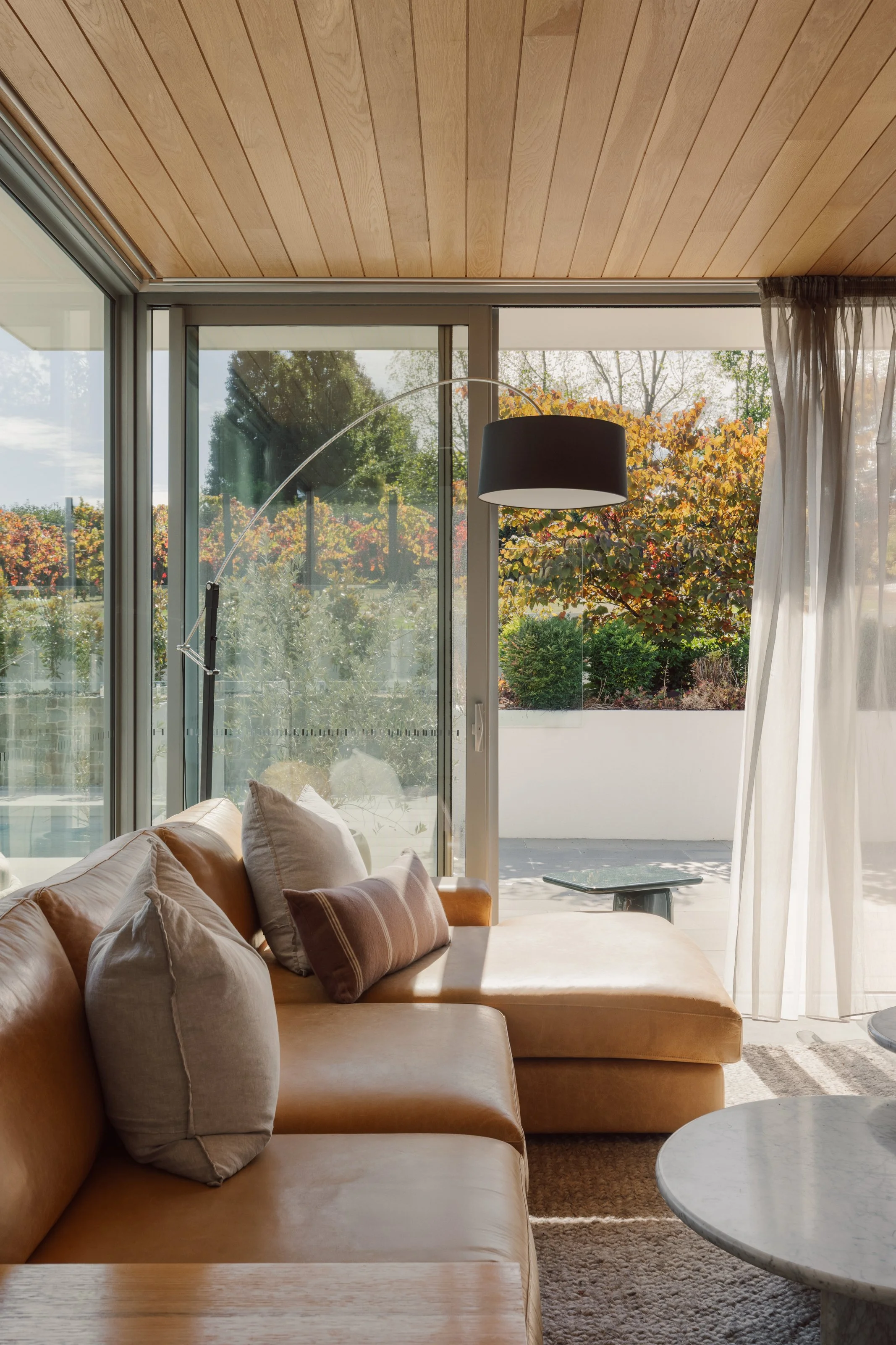 Living room with tan leather sectional sofa and multiple cushions next to large glass sliding door leading to a balcony with outdoor greenery.
