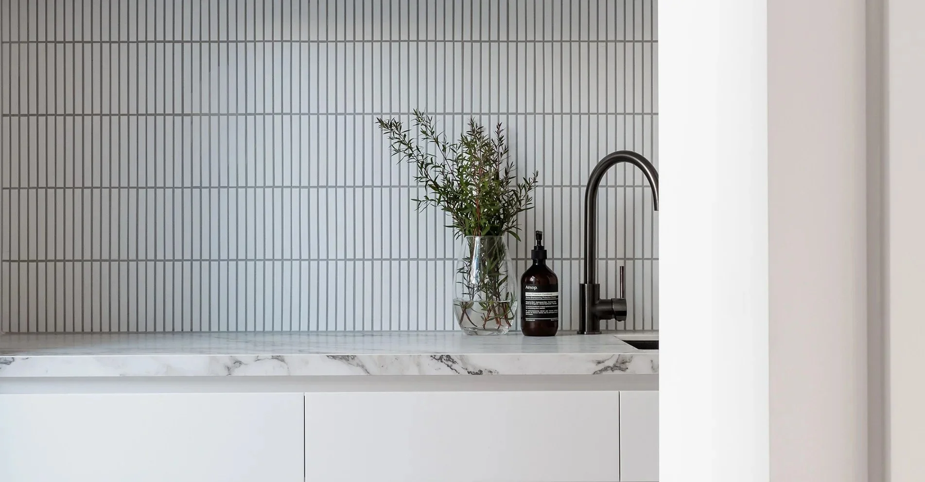 Kitchen countertop with a clear vase of green plant, a black soap dispenser, and a black faucet against a tiled gray backsplash.