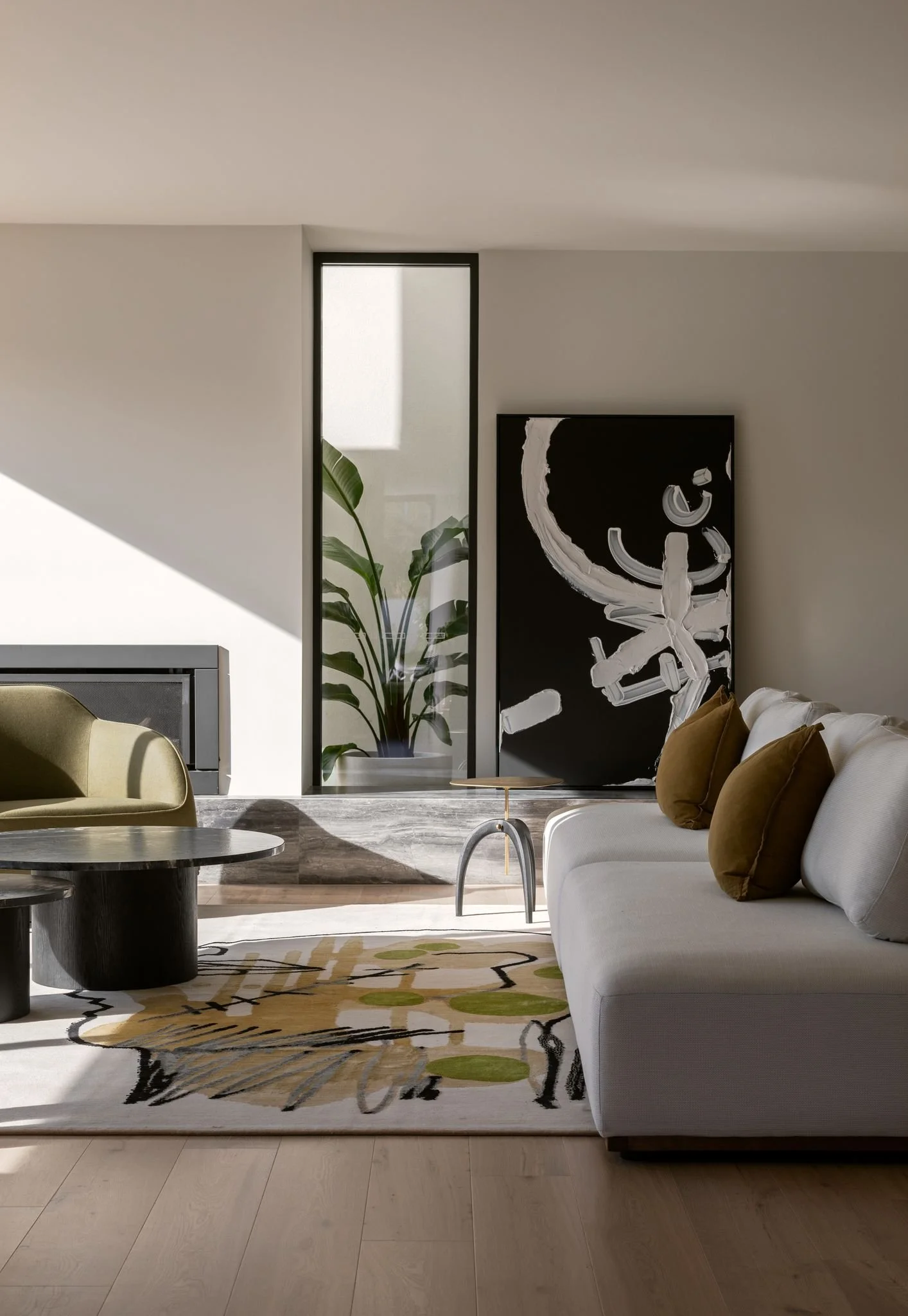 Modern living room with white sofa, brown pillows, abstract black and white artwork, green armchair, glass-paneled corner with large potted plant, patterned area rug, and light wood flooring.