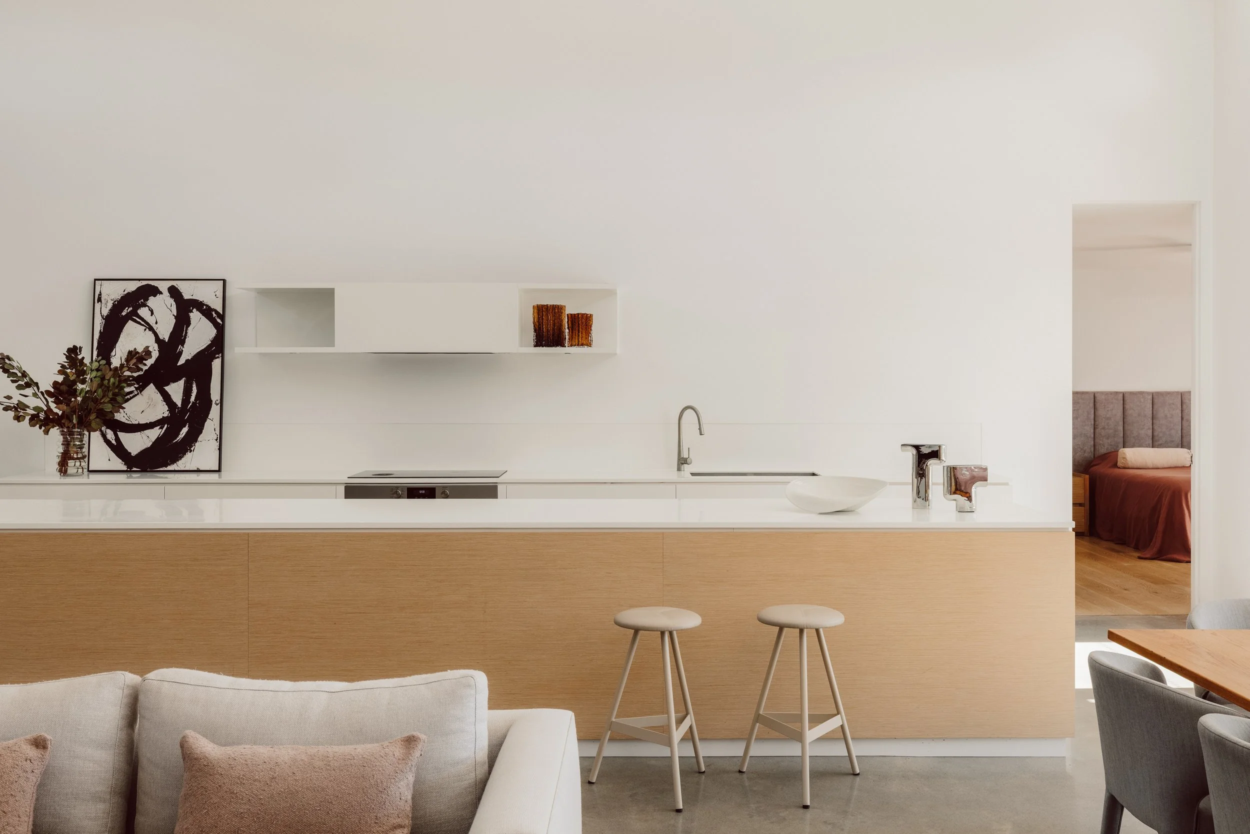 Minimalist kitchen with white walls, a wooden island, two beige barstools, and a glimpse of a bedroom with a bed and headboard in the background.