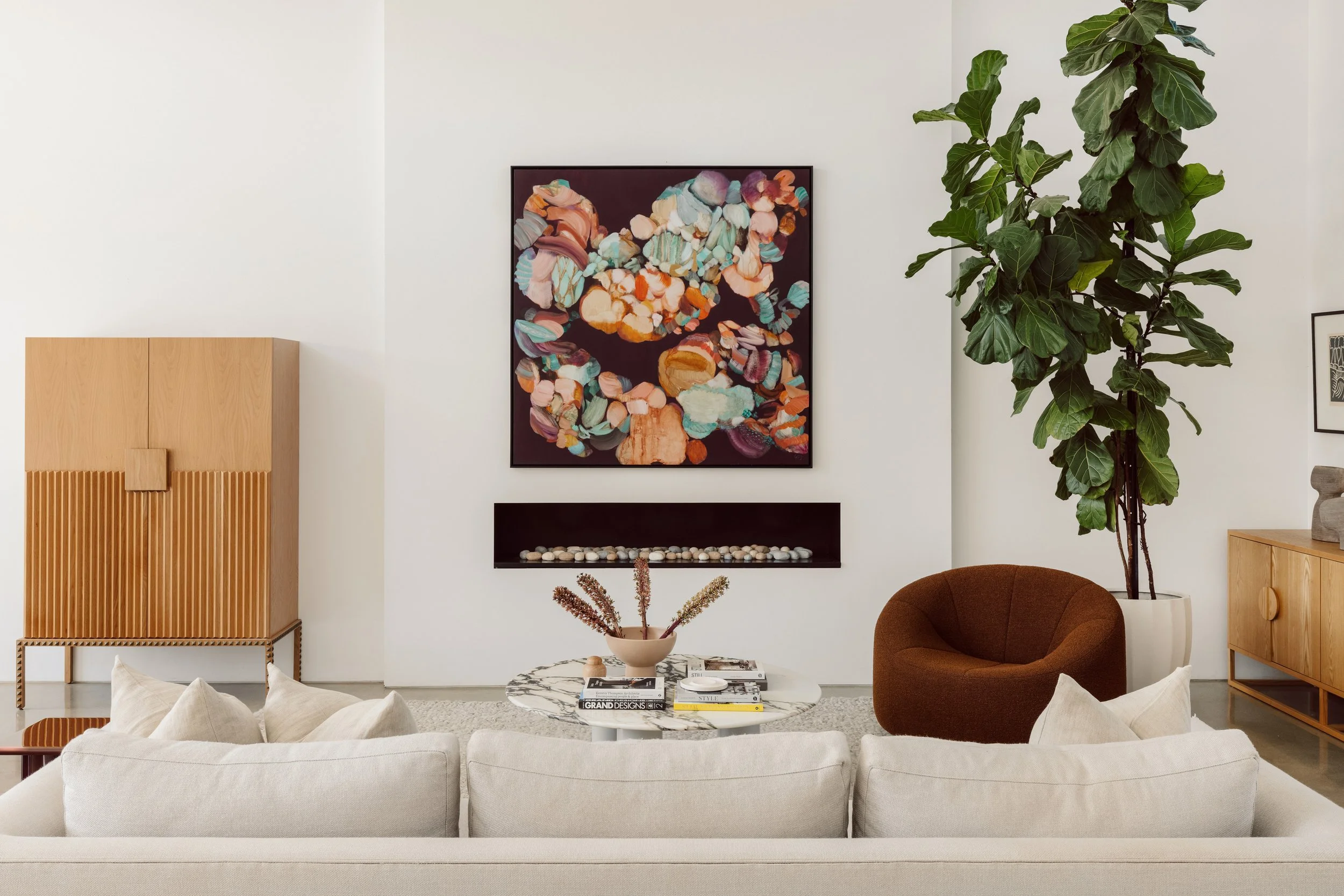 Living room with white walls, a beige sofa, a marble-top coffee table with books and dried flowers, a large green plant, a round brown armchair, a wooden cabinet, artwork on the wall, and a black rectangular wall shelf with decorative stones.