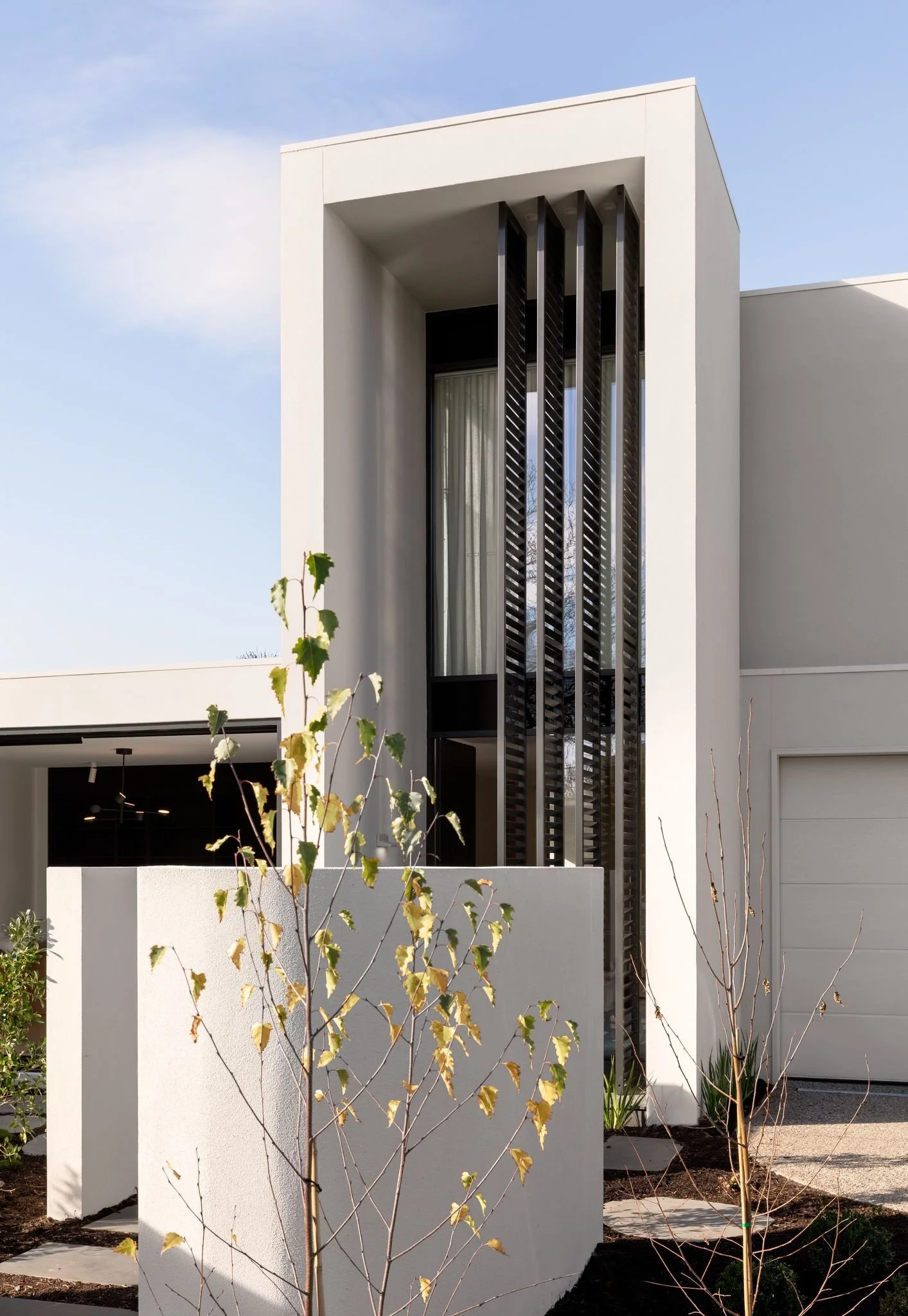 Modern white residential building with large vertical window, black slatted architectural feature, white fence, and sparse young trees in front