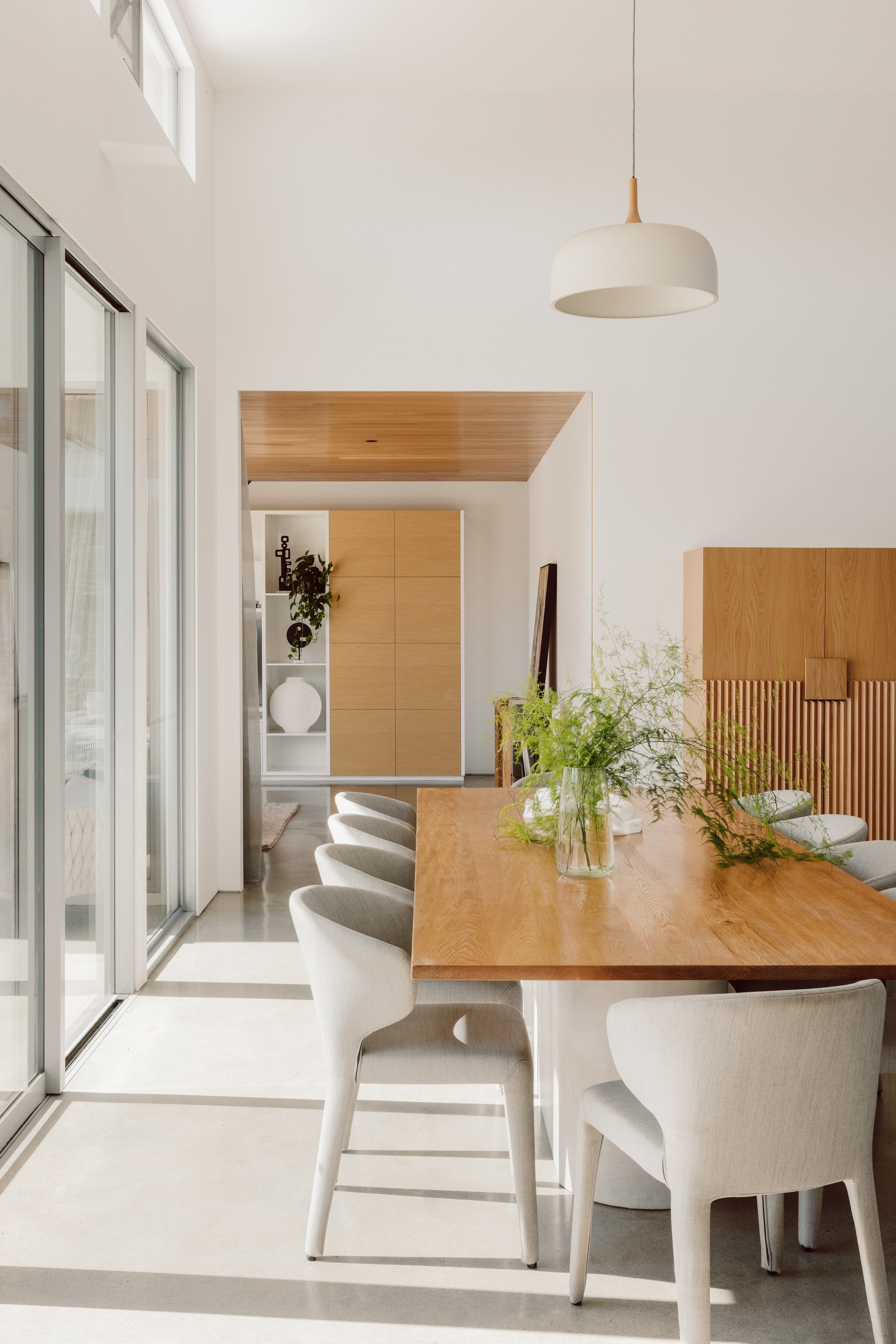 A bright, modern dining room with a large wooden table, white chairs, and a glass vase with greenery. Sunlight streams in through sliding glass doors, and a simple white pendant lamp hangs from the ceiling.