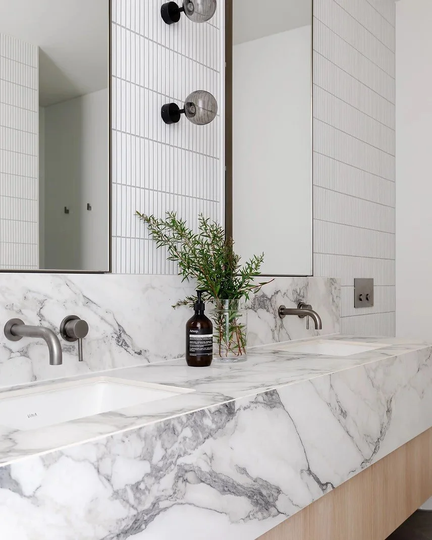 Modern bathroom vanity with two marble sinks, metallic faucets, a large mirror, a brown bottle with green foliage, and a wall-mounted light fixture.
