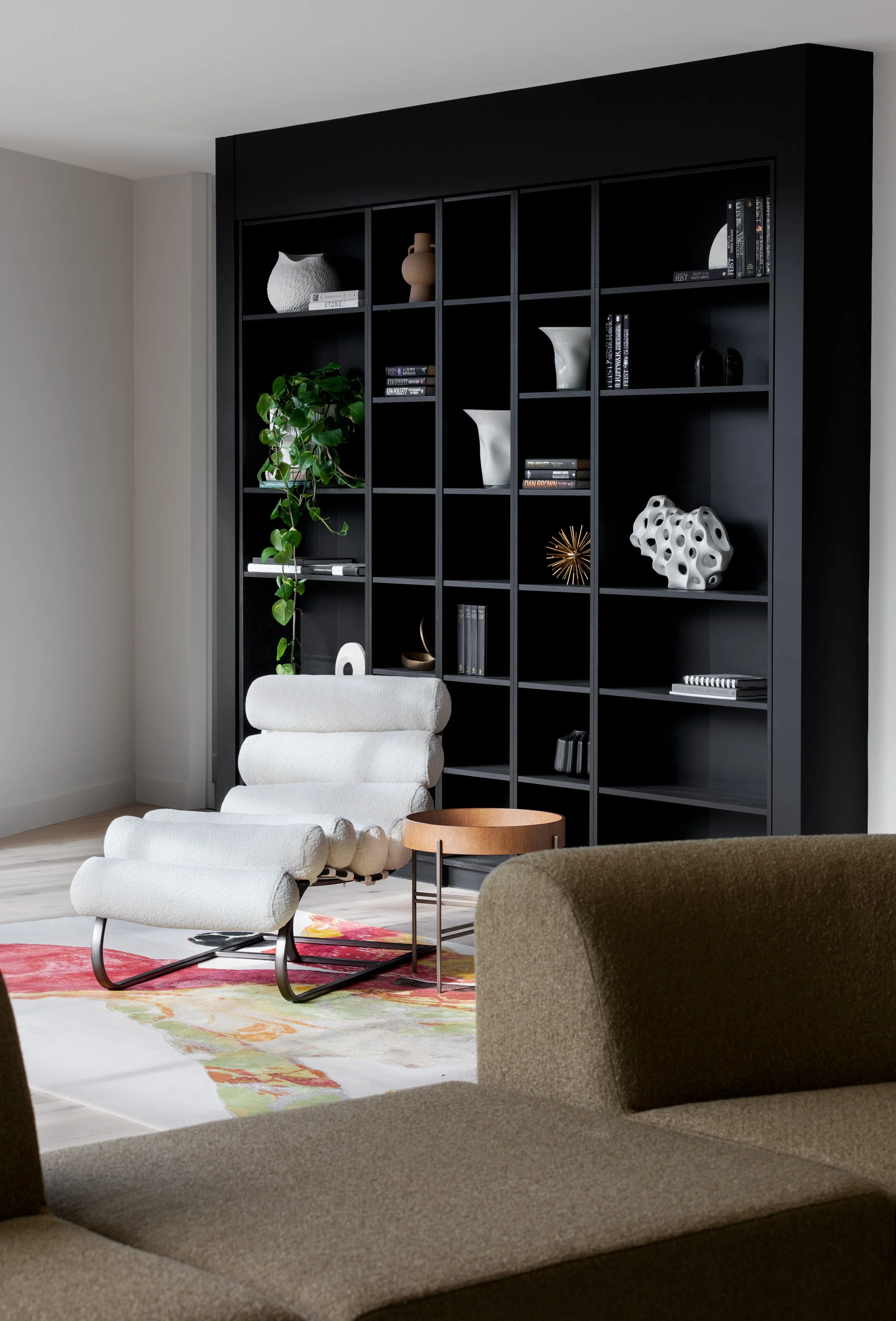 Modern living room with a black bookshelf filled with decorative objects, books, and a green plant. There is a white lounge chair with a matching ottoman, a round wooden side table, and part of a beige sofa visible. A colorful area rug is on the light wood floor.