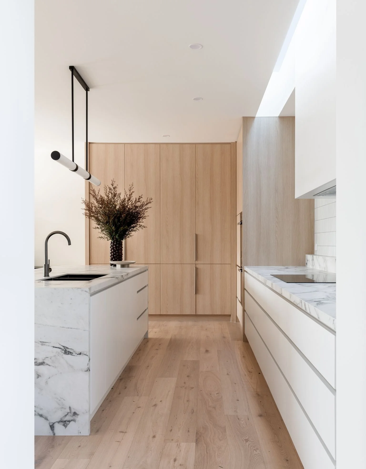 Modern minimalist kitchen with white and marble countertops, light wood cabinets, and a black pendant light fixture.