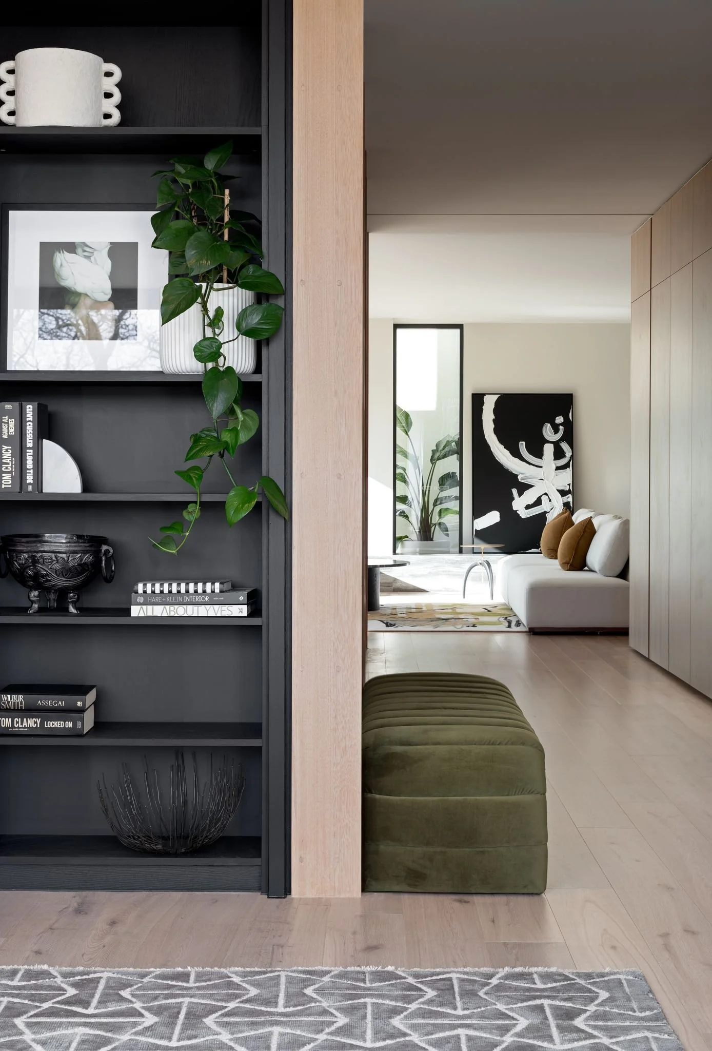 Interior view of a modern living room with a black bookshelf, green plant, and a sofa with brown and white pillows, complemented by modern artwork and tall plants in the background.