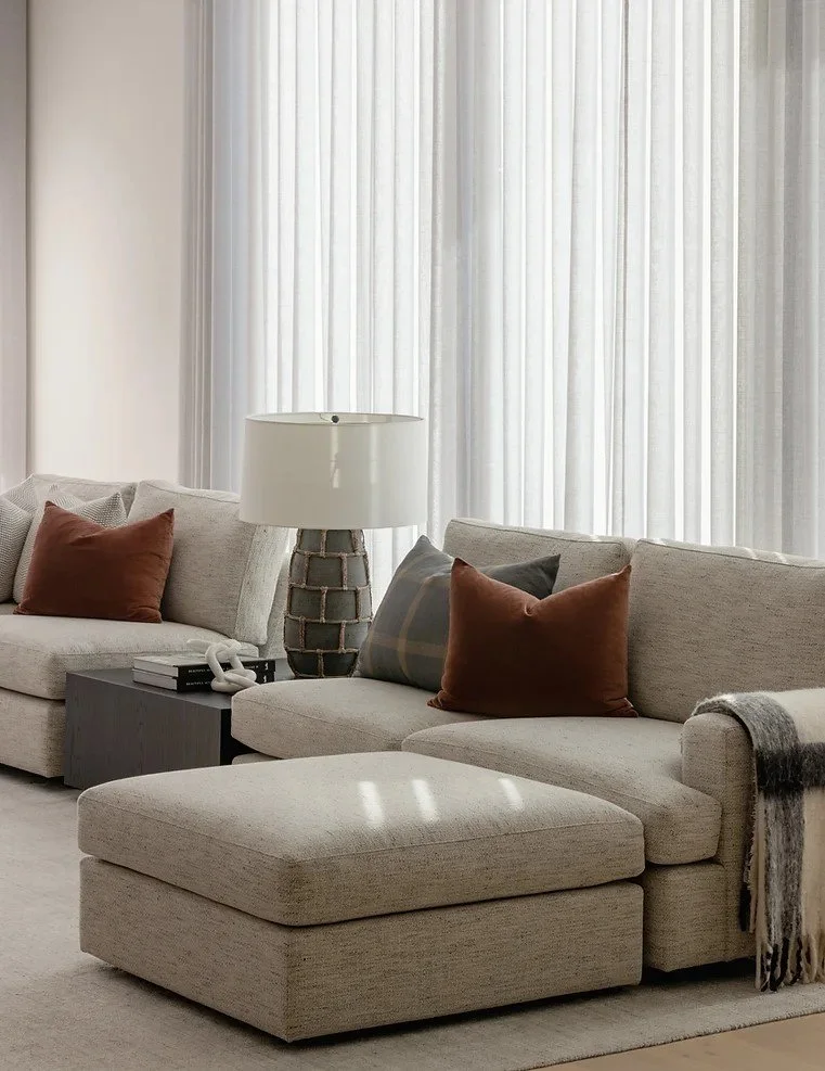 A living room with a beige sectional sofa, decorative pillows, a large table lamp, and a dark side table with books and decor, with sheer white curtains in the background.