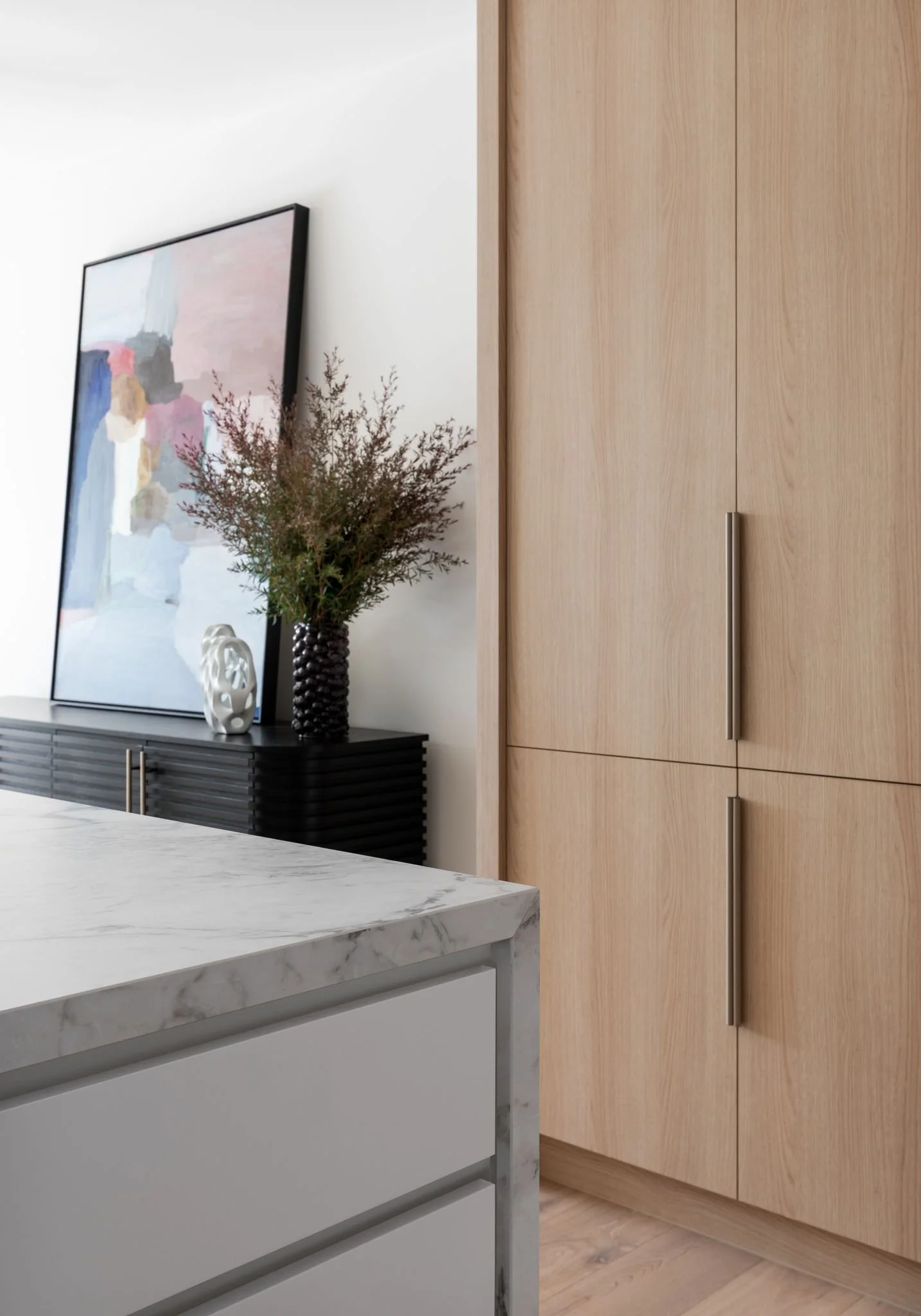 Part of a modern kitchen or living area with a white marble countertop, light wooden cabinets, a black sideboard with decorative items, and an abstract painting on the wall.