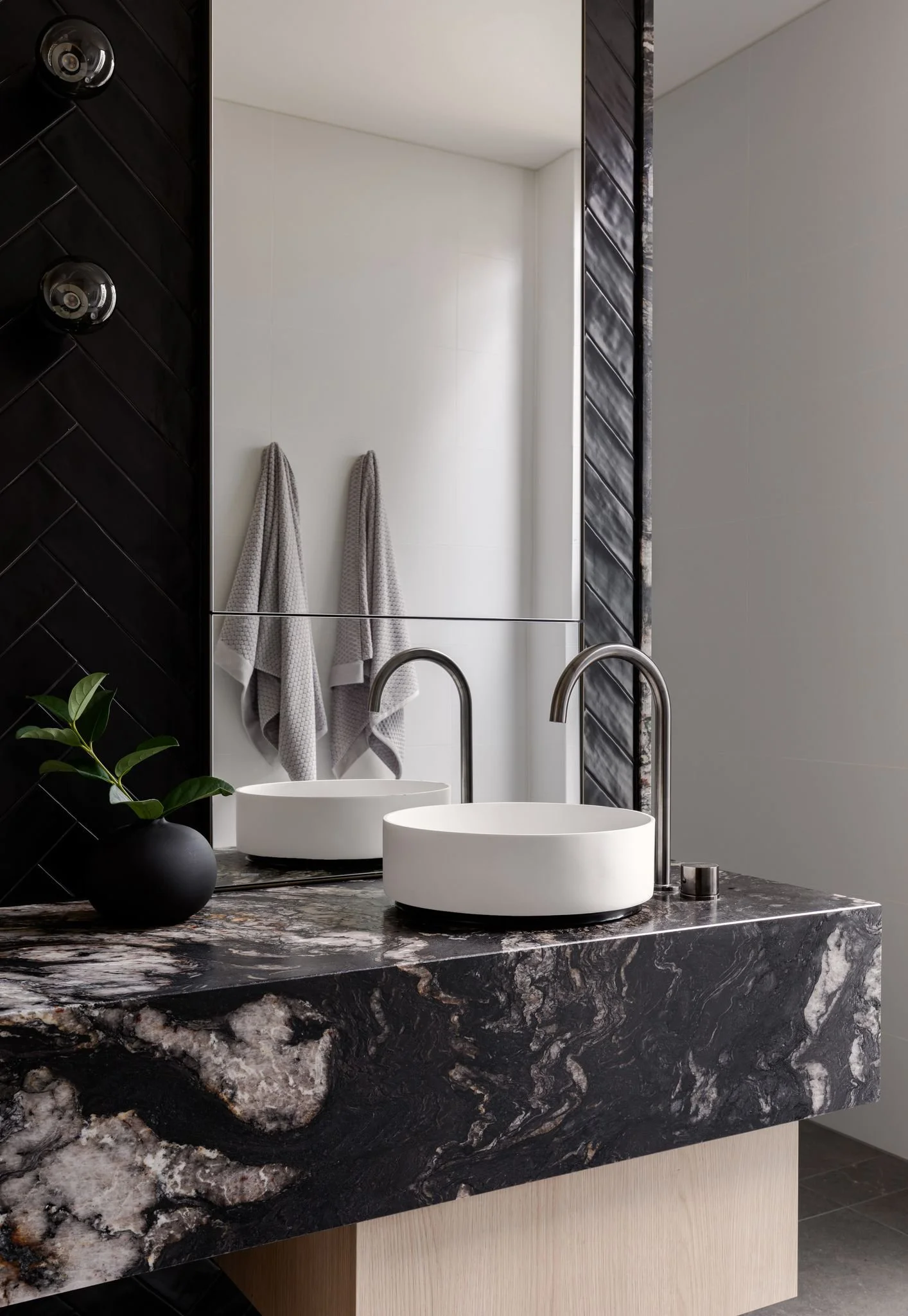 Modern bathroom with a black and white marble countertop, white round vessel sinks, and a large mirror. Two gray towels hanging on the wall and a small black vase with green leaves on the counter.