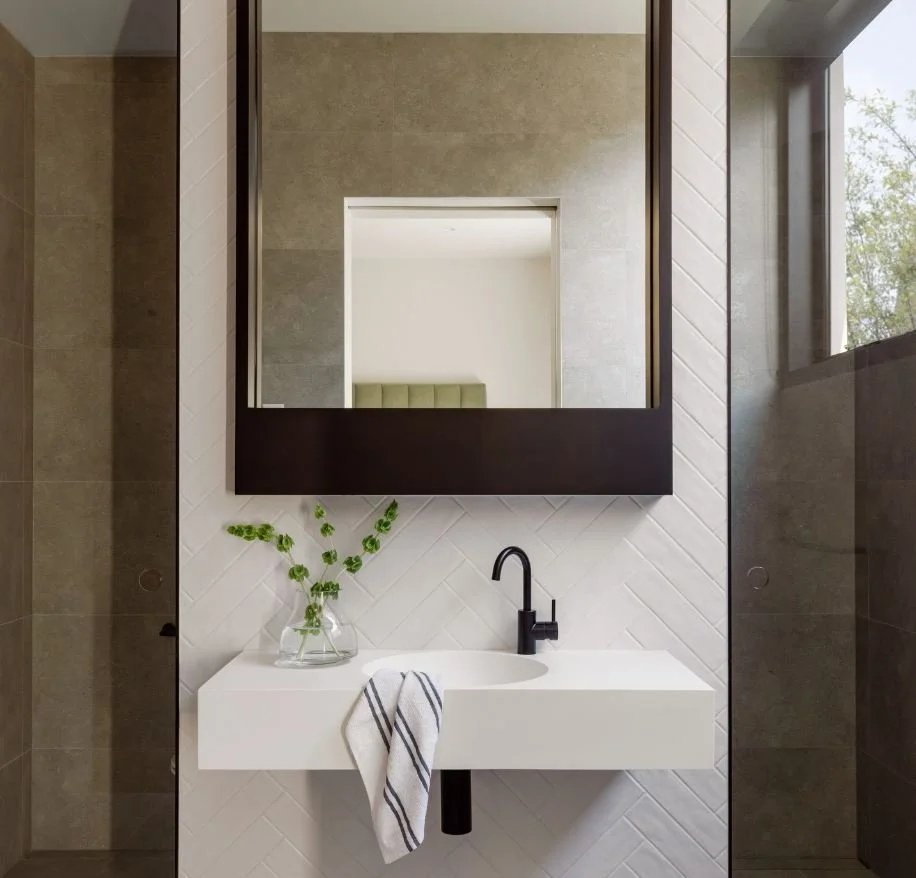 Modern bathroom with a white rectangular sink, a black faucet, a decorative glass vase with green foliage, a large mirror, gray tiled walls, and a window with a view of trees.