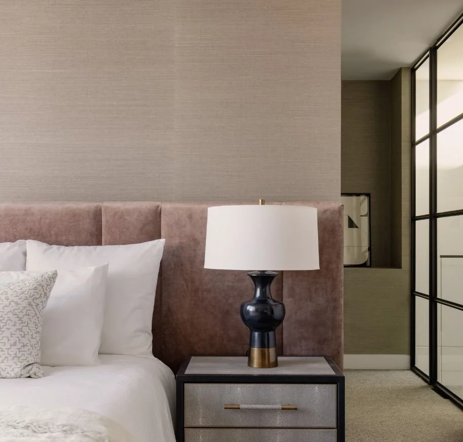 Modern bedroom with a beige textured wall, pink upholstered headboard, white bedding, decorative pillows, black and gold table lamp, and a nightstand.