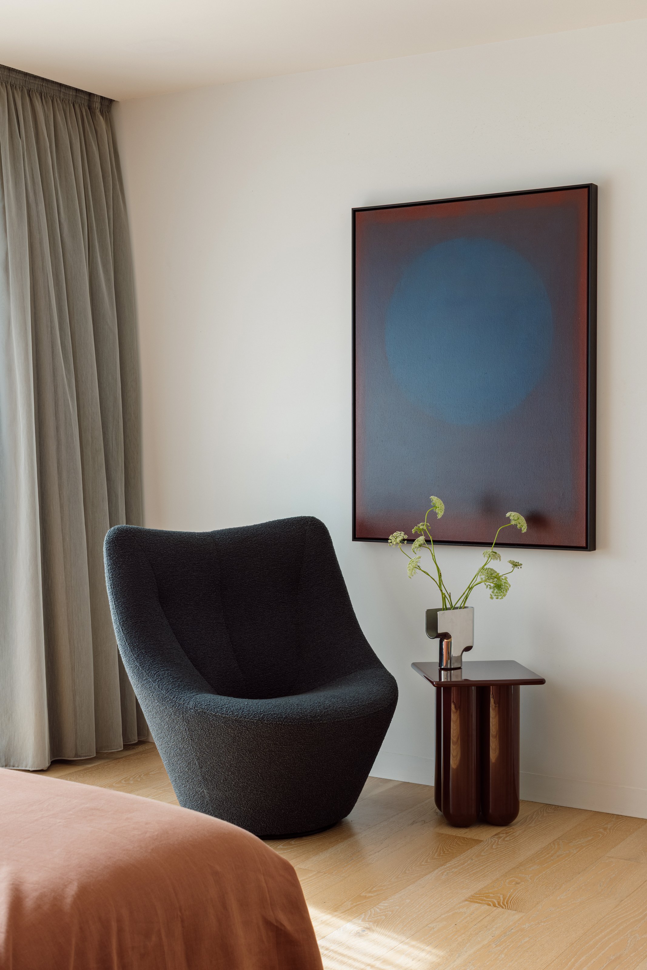 A modern bedroom corner with a black textured armchair, a small wooden side table with a vase of green flowers, a large abstract painting with a blue circle on a red background, beige curtains, and a bed with a rust-colored blanket.