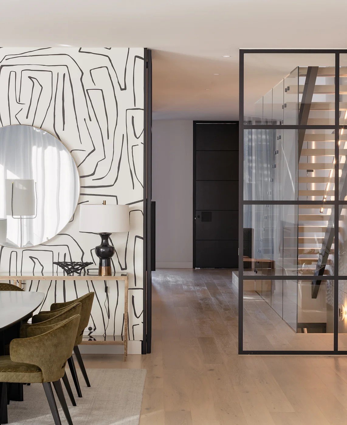 Modern dining area with a black and gold table, olive green chairs, a wall with black abstract line art, a large round mirror, a zebra design table lamp, a decorative black and gold table, and a glass staircase enclosed in a black metal frame.