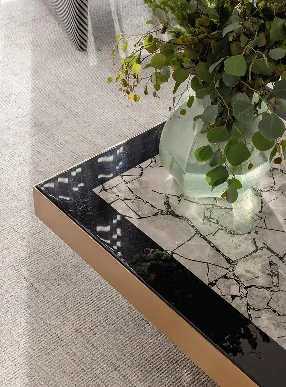 Close-up of a black marble-topped coffee table with a mosaic pattern, holding a large green ceramic vase with a leafy green plant, situated on a beige textured carpet