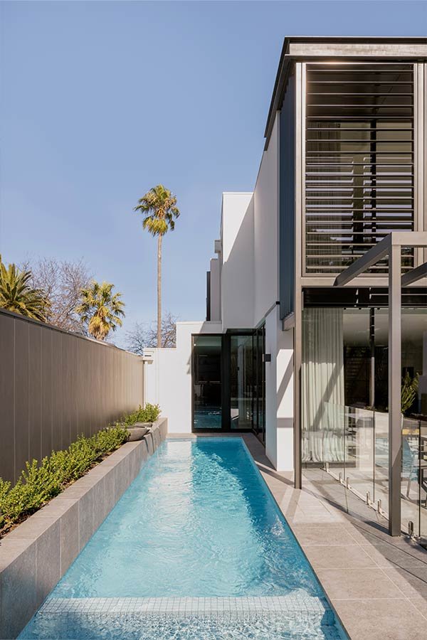 Modern house with a narrow swimming pool, glass sliding doors, and palm trees in the background under a clear blue sky.
