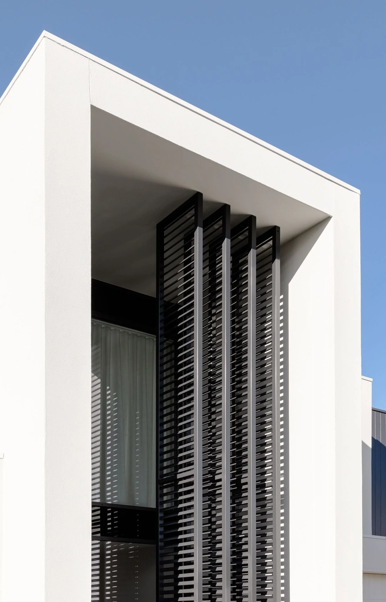 Close-up of a modern white building facade with black vertical slats and a large window under a clear blue sky.