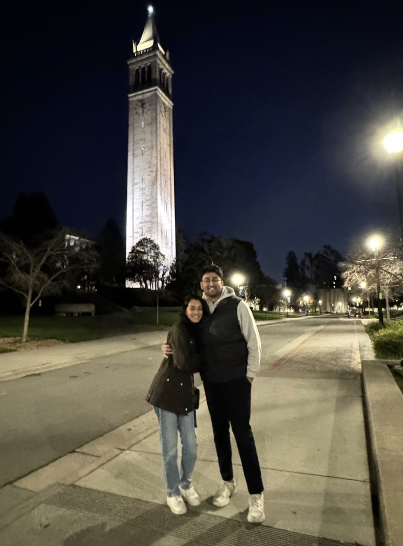 A couple standing close together on a sidewalk at night, smiling with the illuminated Big Ben clock tower in the background.