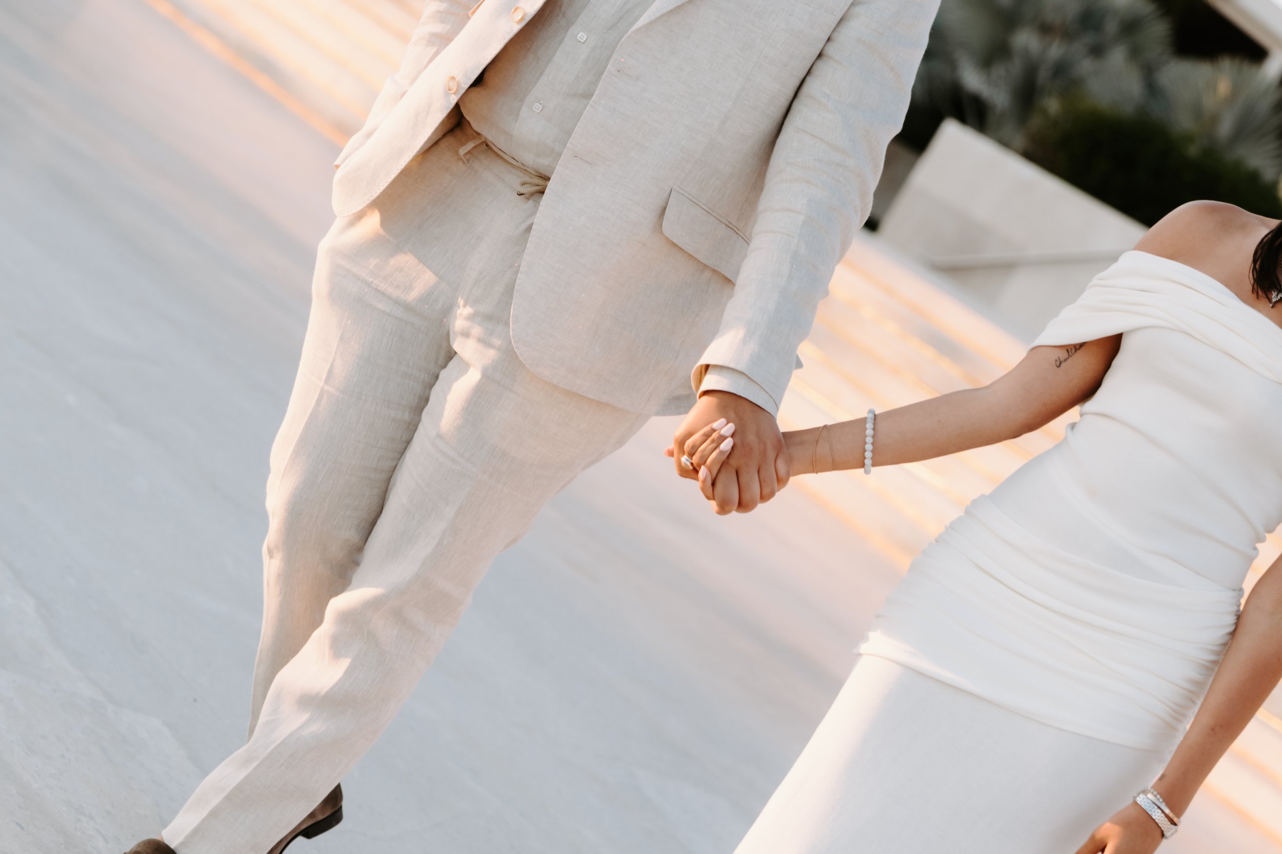 Close-up of a couple holding hands, the woman in a white dress and the man in a light-colored suit, with a sunset background.
