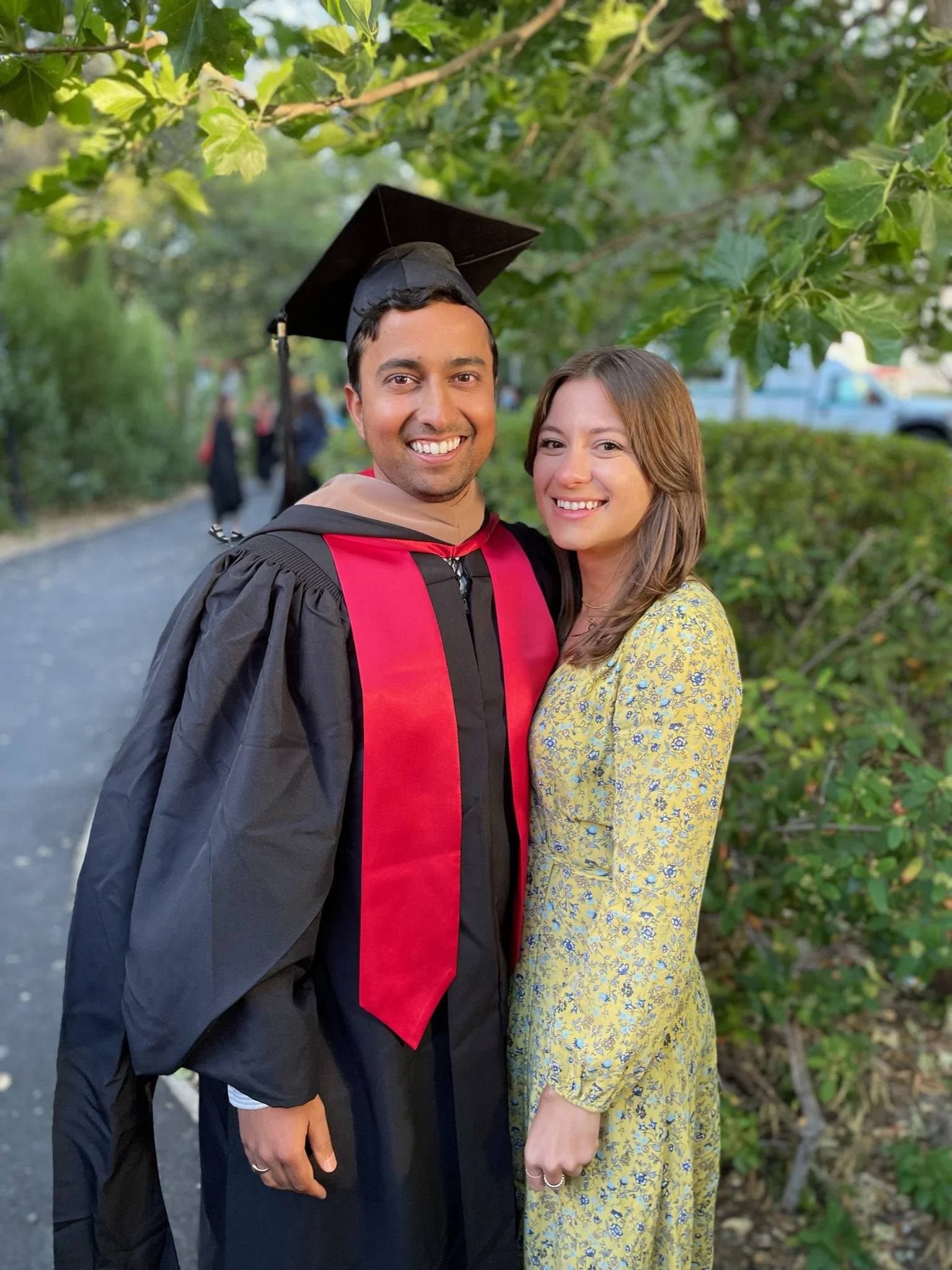 A young man in a graduation gown and cap standing close to a young woman in a yellow floral dress outdoors under green trees.