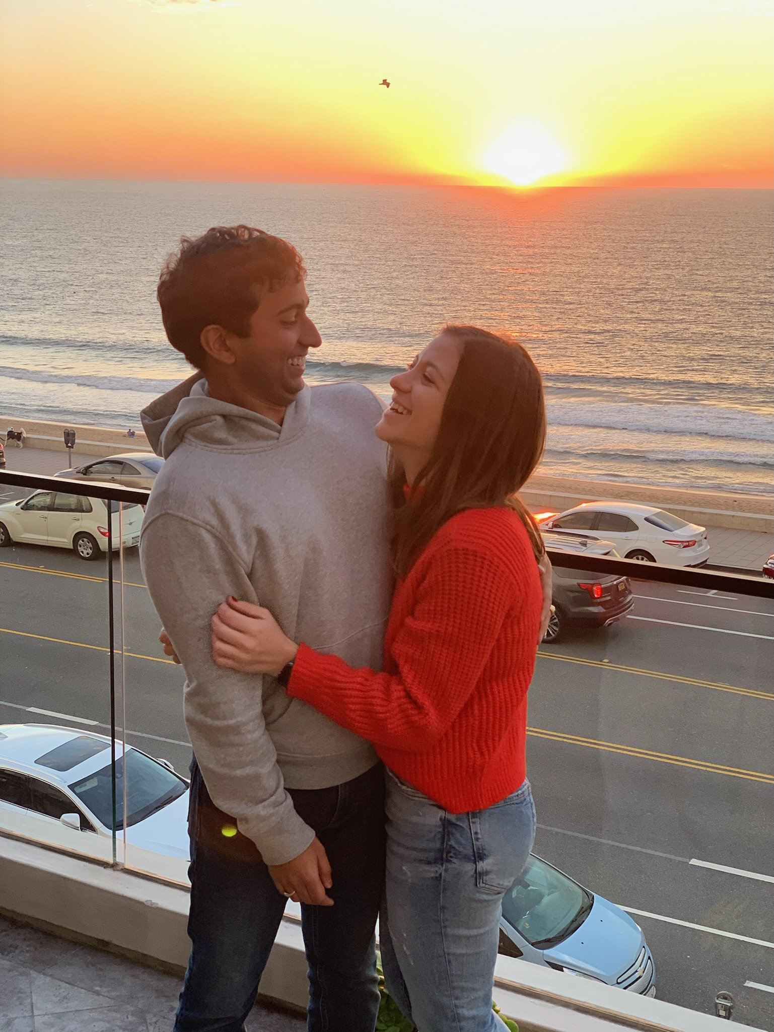 A smiling couple stands on a balcony overlooking the ocean at sunset, embracing each other happily.