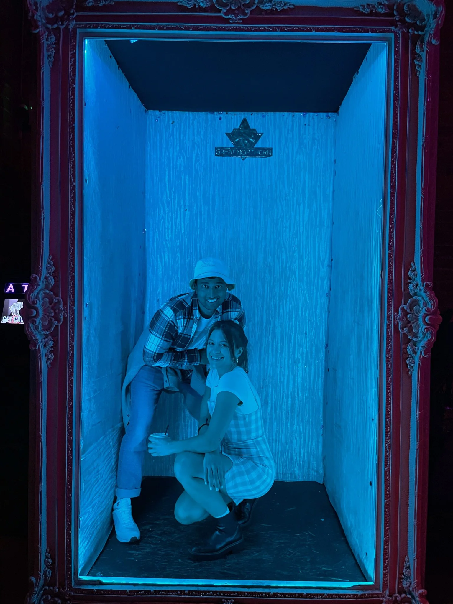 Two people, a man and a woman, posing inside a small, illuminated photo booth with blue lighting. The man is standing and wearing a white bucket hat, a plaid shirt, light-colored pants, and white shoes, while the woman is kneeling with one knee on th