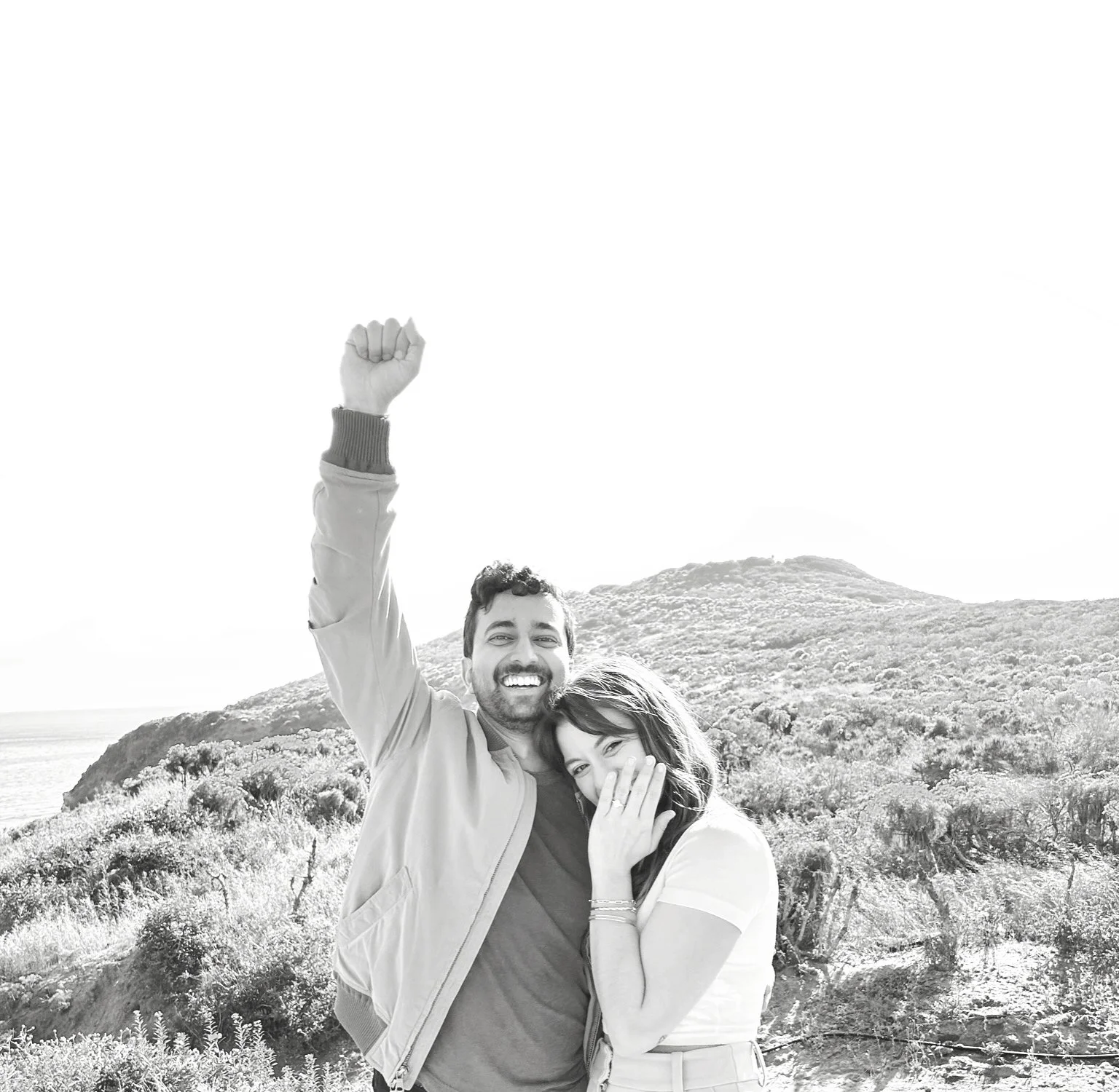 Black and white photo of a happy couple outdoors, with the man raising his arm in celebration and the woman smiling and covering her mouth.