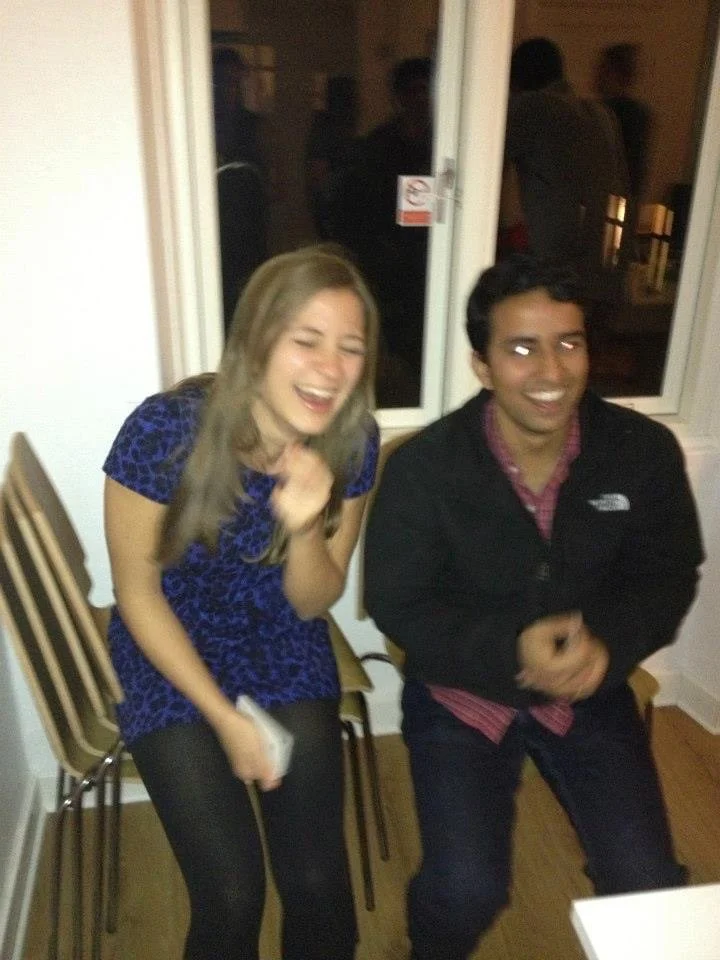 Two young adults laughing and enjoying themselves, sitting on chairs near a window at a social gathering.