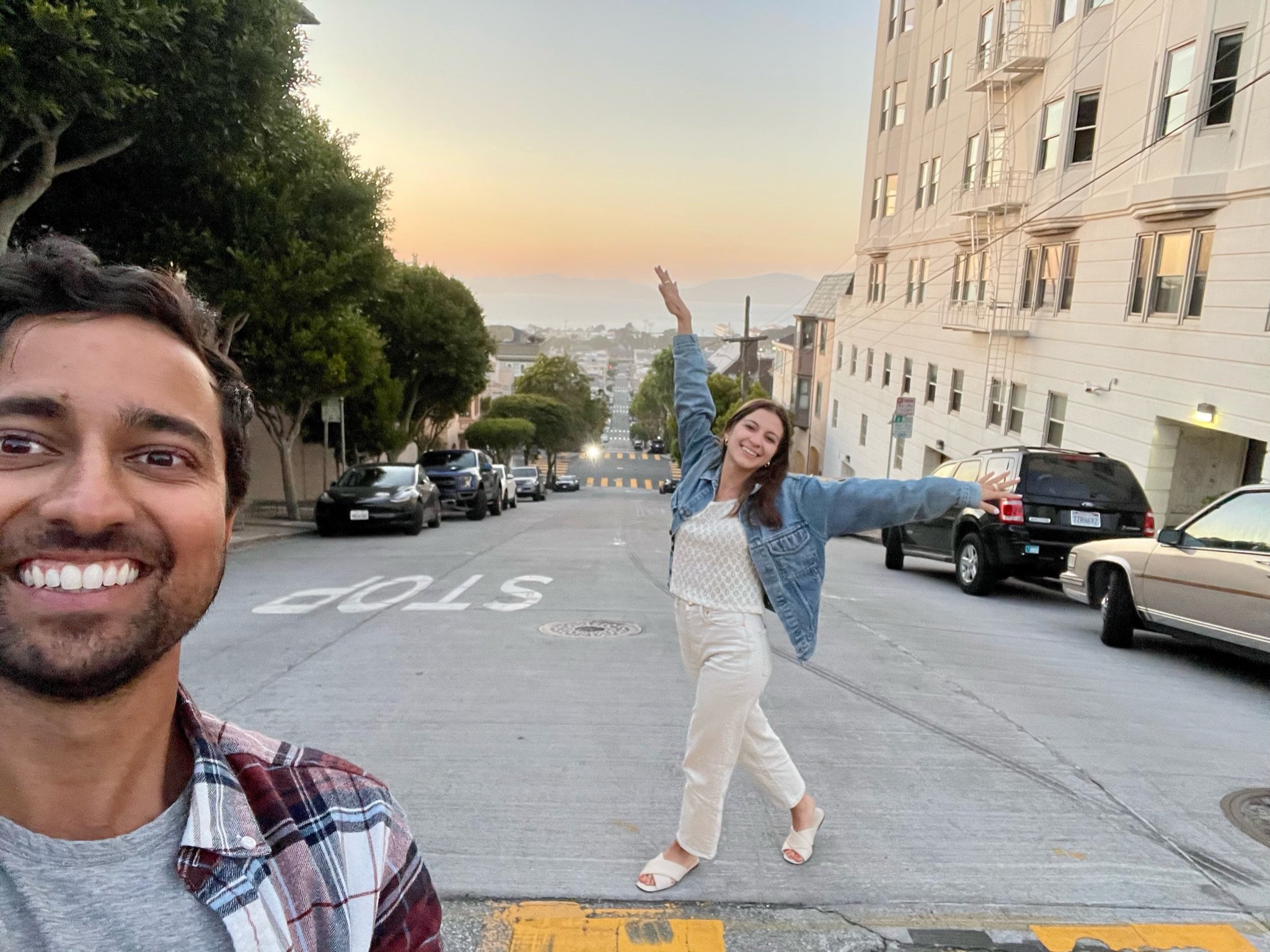 A man and a woman smiling on a city street at sunset. The woman is in the background, standing on one leg with her arms outstretched, wearing a denim jacket and white pants. The man is in the foreground, taking a selfie, wearing a plaid shirt and a g