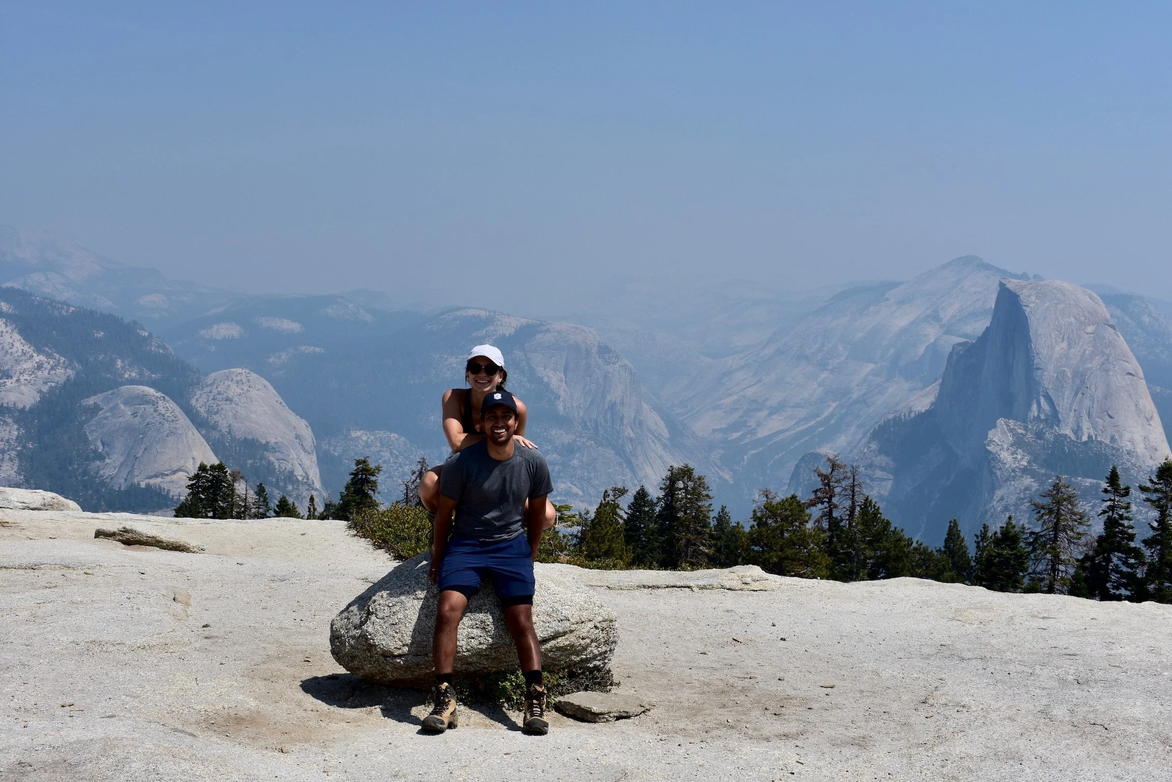 Two people, a woman and a man, smiling and posing on a large rock with mountains and trees in the background.
