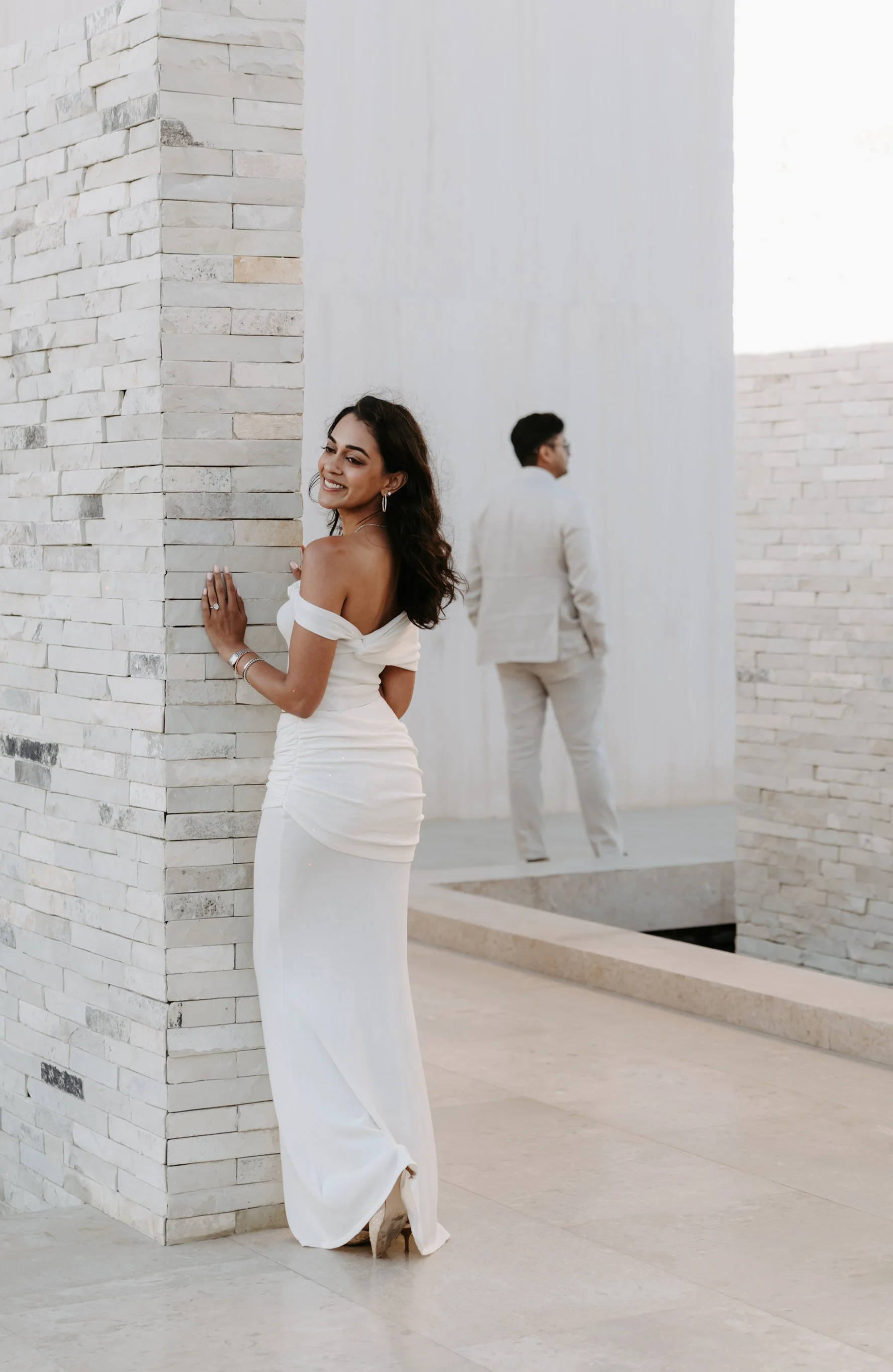 A woman in a white dress smiling and leaning against a brick wall, while a man in a beige suit stands in the background with his back to the camera.