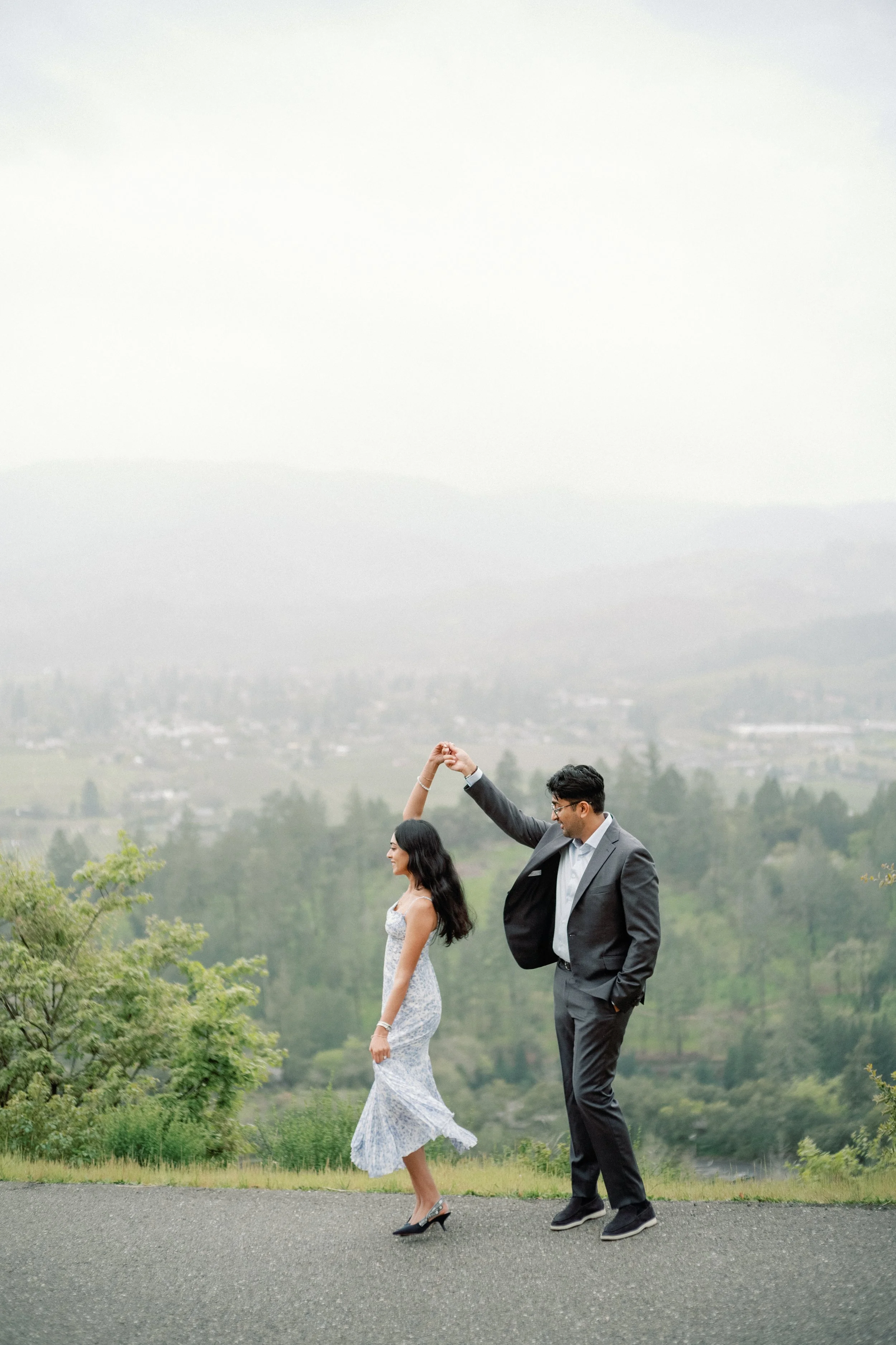 A couple dancing outdoors on a gray paved surface with a mountain and forest background. The woman wears a white and blue floral dress and black heels, and the man is in a gray suit with a white shirt and black shoes. He is holding her hand as they d
