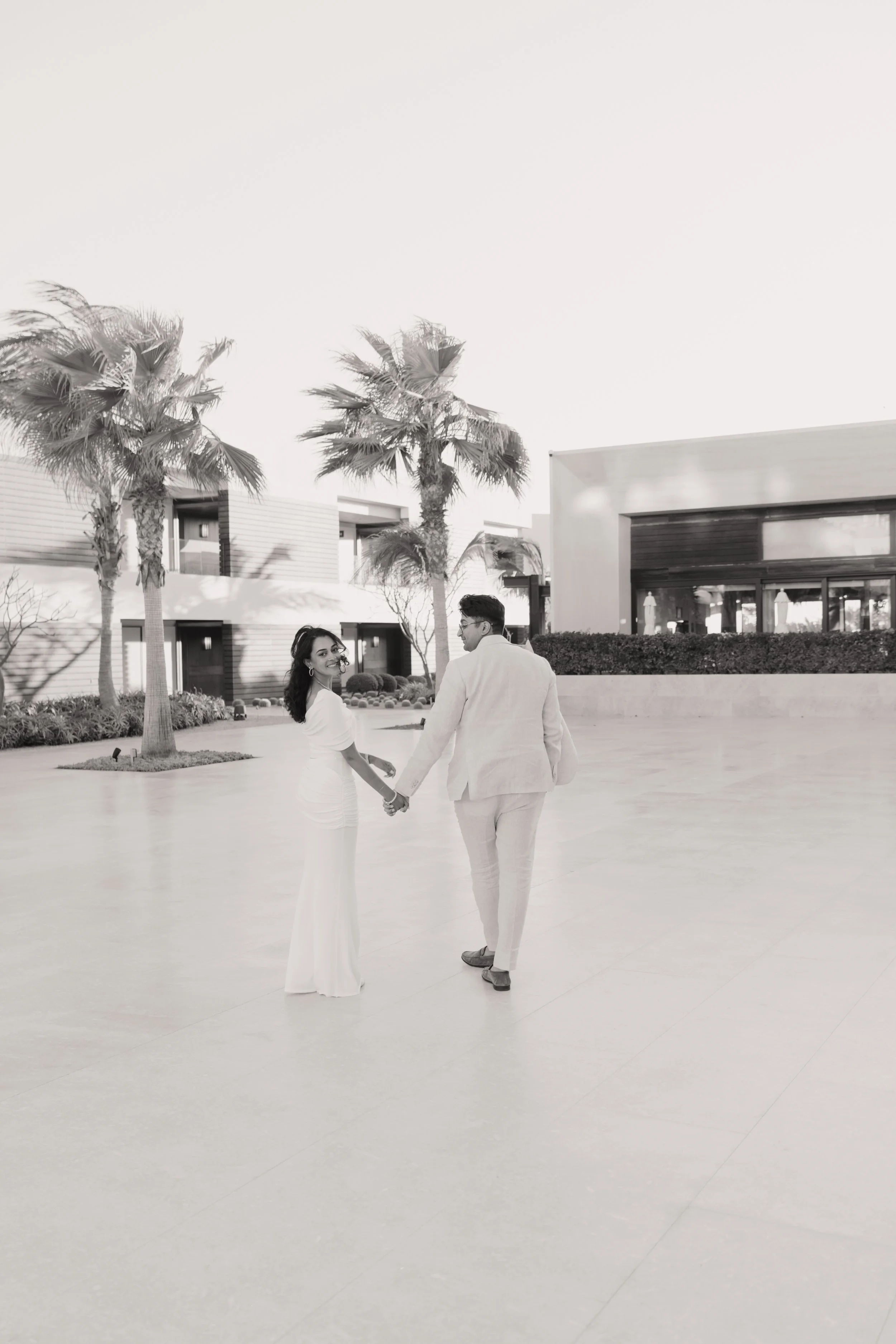 A couple holding hands and looking at each other in an outdoor courtyard with palm trees and modern buildings in the background.