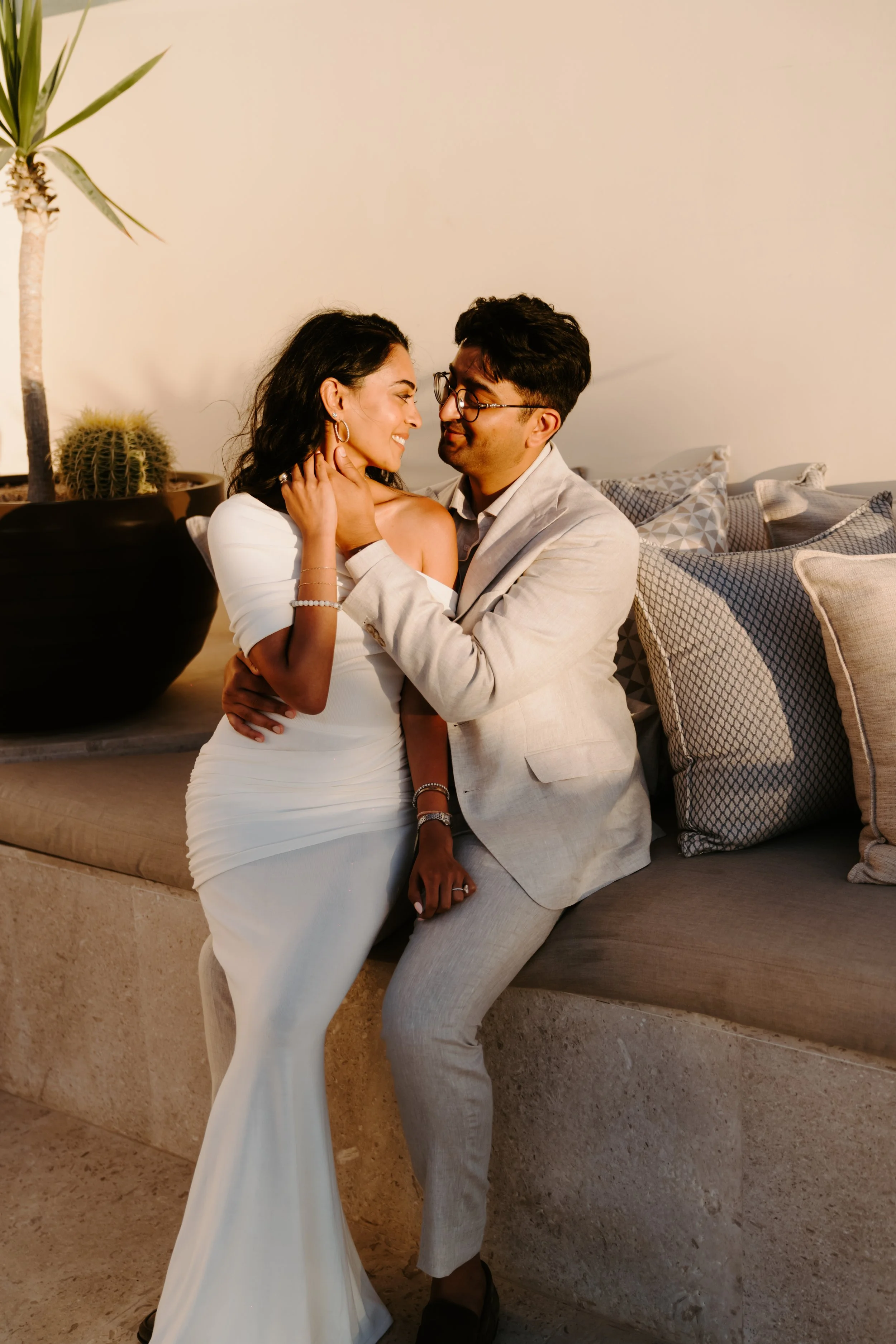 A couple dressed in formal attire sharing a romantic moment indoors, with the woman in a white gown and the man in a light-colored suit, near decorative pillows and plants.