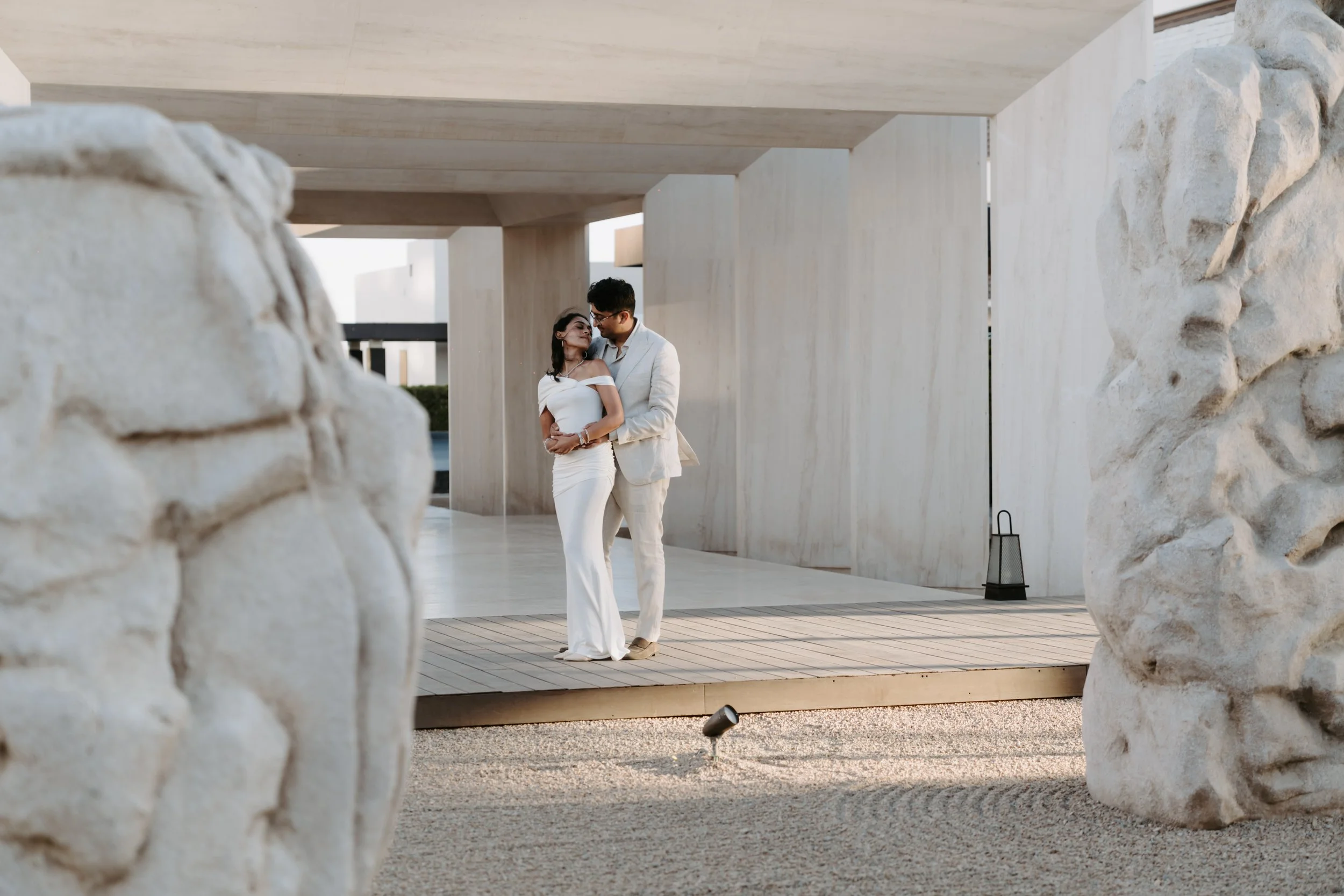 A couple dressed in white clothing dancing closely under a modern architectural structure with large stone accents.